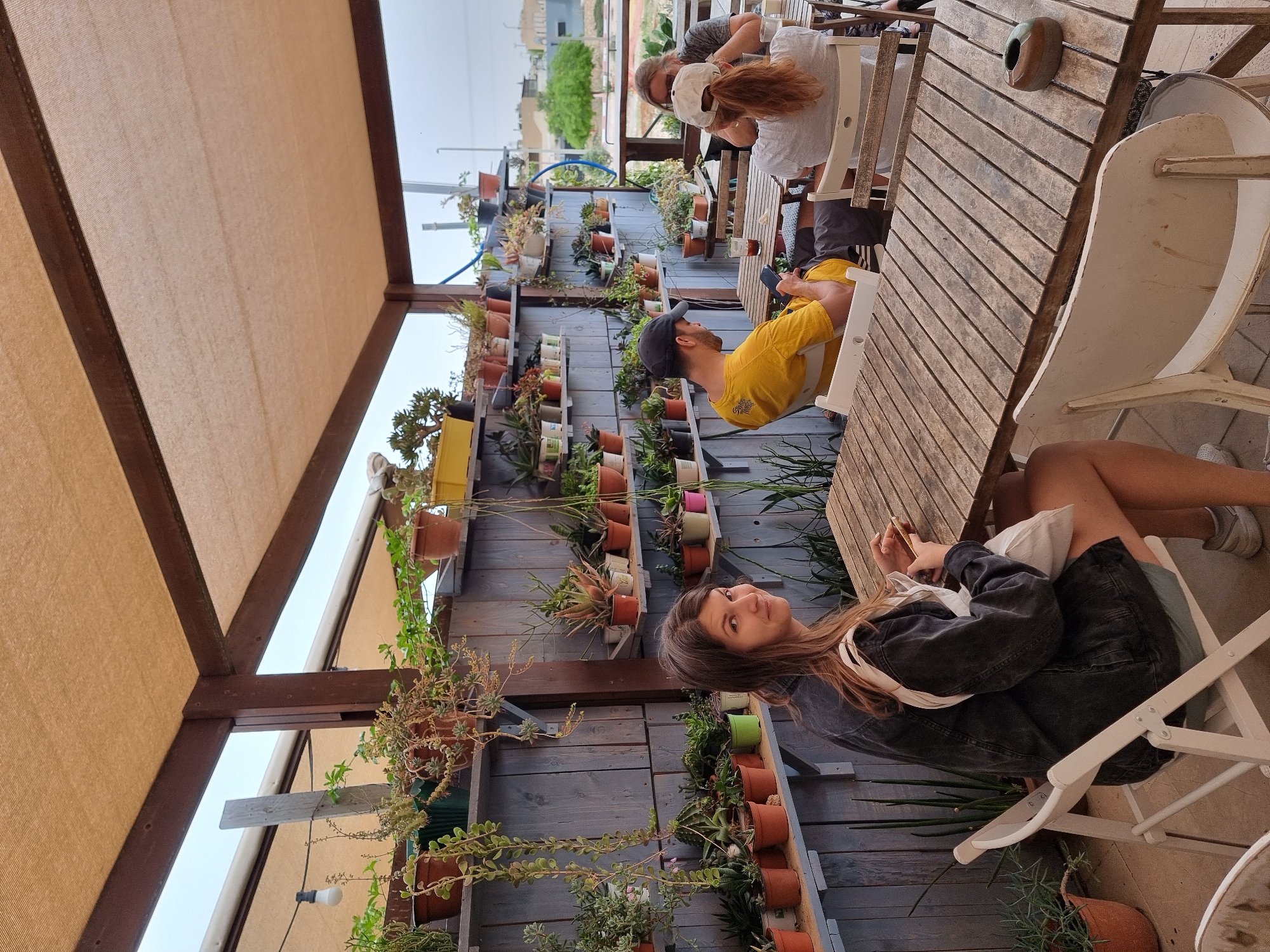 The image captures a scene in Mitzpe Ramon, Israel, featuring several people at a wooden table and chairs. The location appears to be an outdoor patio area with a shade covering. A wall behind the table is adorned with numerous potted plants arranged in rows.
One woman is seated at the table, looking directly at the camera, wearing a dark denim jacket. Further down the table, a man in a yellow shirt and a baseball cap sits facing away from the camera. Two other individuals are visible at the end of the table, appearing to be engaged in conversation. The table is made of wooden slats and holds a brown container.
The surroundings suggest a relaxed, casual setting with an emphasis on greenery. The lighting indicates either a late afternoon or early morning timeframe. The city of Mitzpe Ramon is implied by the contextual elements of the scene.