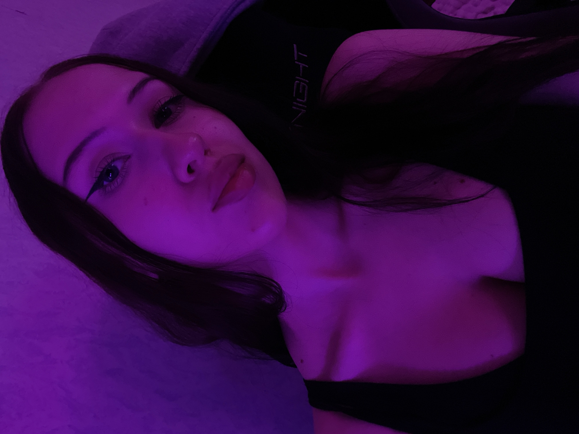 A young woman is captured in a close-up, selfie-style shot. She has long, dark hair and is wearing a black tank top. The lighting is predominantly purple, creating a moody and atmospheric effect. She is leaning against what appears to be a gaming chair, with the brand name "NIGHT" visible on it. The background is a textured wall, also illuminated by the purple light. Her makeup includes winged eyeliner. The overall impression is one of a casual, indoor setting, possibly a bedroom or a gaming setup. There is no information in the image that would allow for the determination of its location in Votkinsk, Russia.