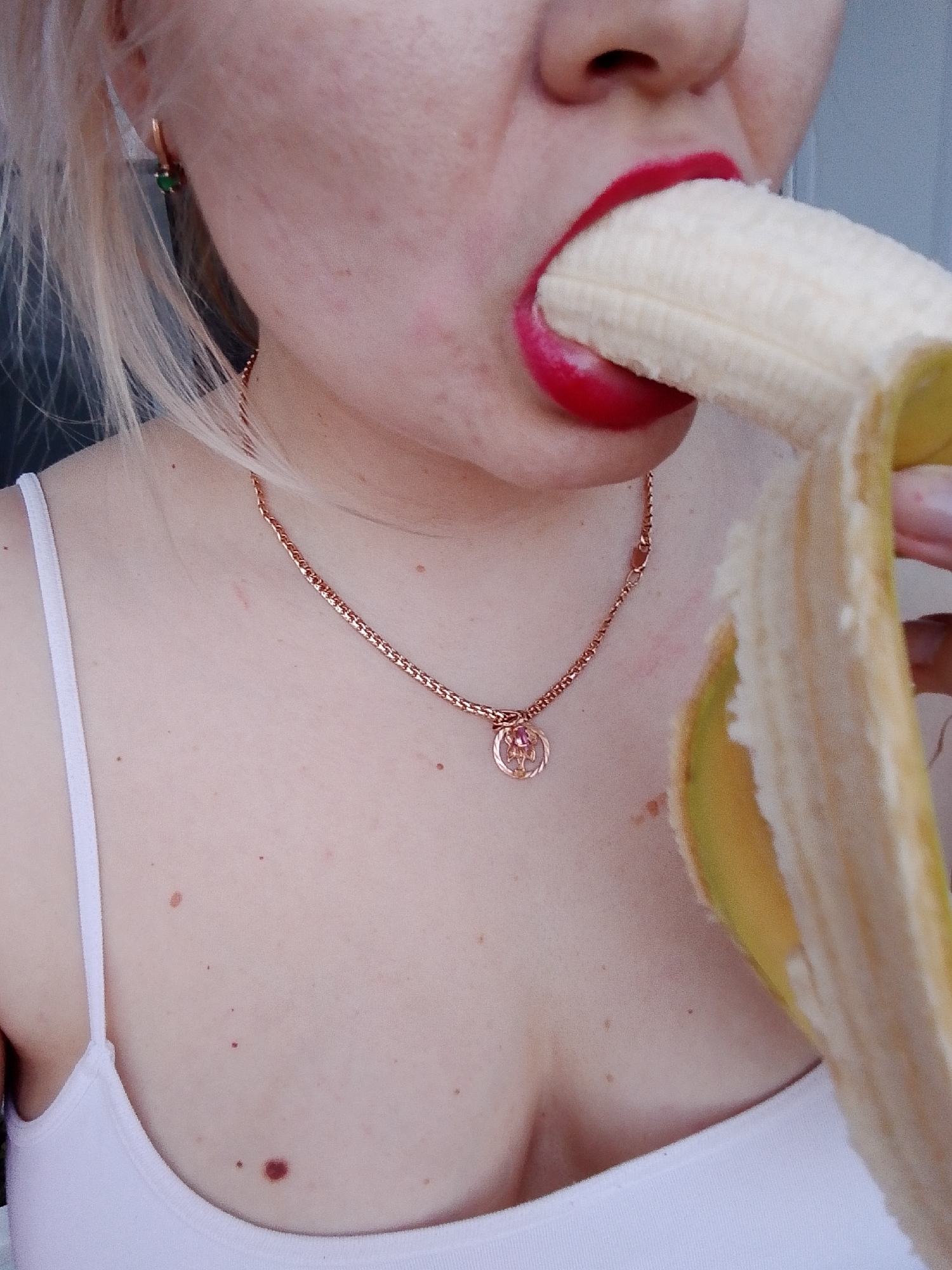 A close-up photograph shows a fair-skinned person with blond hair eating a peeled banana. The person has red lipstick on their lips. They are wearing a white tank top and a rose gold necklace with a round pendant. A small green earring is visible in their left ear. The background is out of focus and appears to be a light gray surface. There is no evidence to suggest this photo was taken in Izhevsk, Russia.