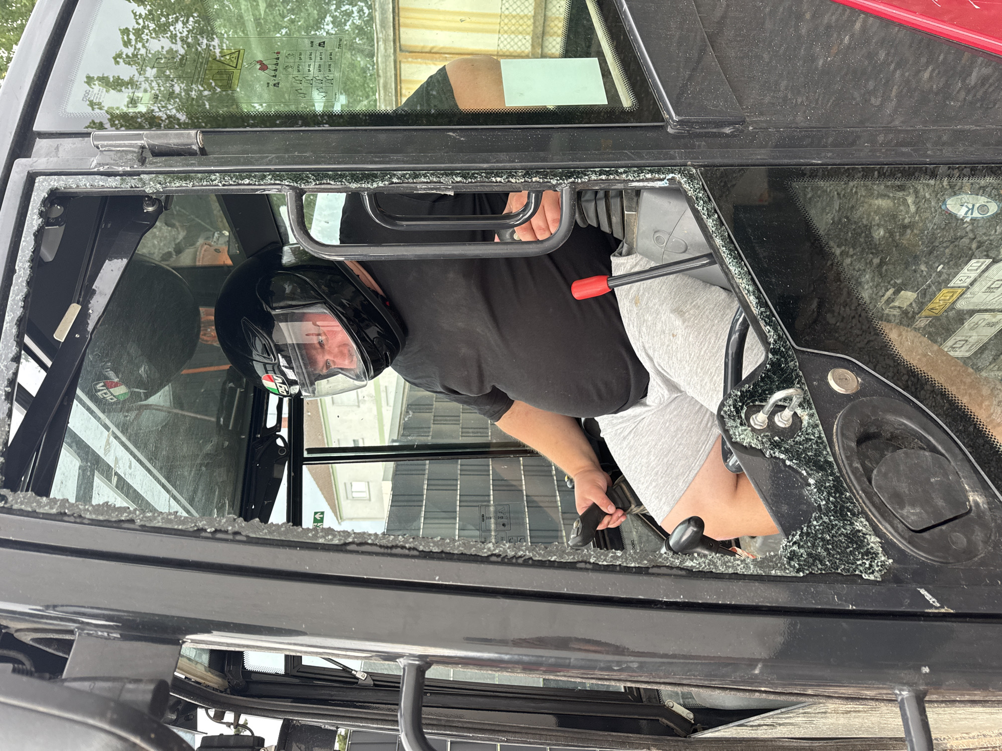 A man wearing a black full-face helmet sits inside the cab of a construction vehicle. The left side window of the cab is broken, with shattered glass visible along the frame and on the floor. The man is wearing a black t-shirt and light grey shorts. He is holding a black joystick control with his right hand. The background shows a building with a white facade and a grey tiled roof, trees, and a fence. The scene is outdoors, suggesting it is located within the city of Bellheim, Germany.