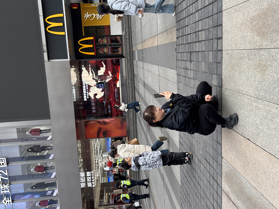 A woman is crouched on a paved walkway, looking at her phone. She is dressed in black pants and a black jacket. Behind her, people are walking past a shop front displaying clothing mannequins, and a McDonald's restaurant. The shop's signage includes Chinese characters and the words "McCafé". There are also some uniformed individuals near the shop front.