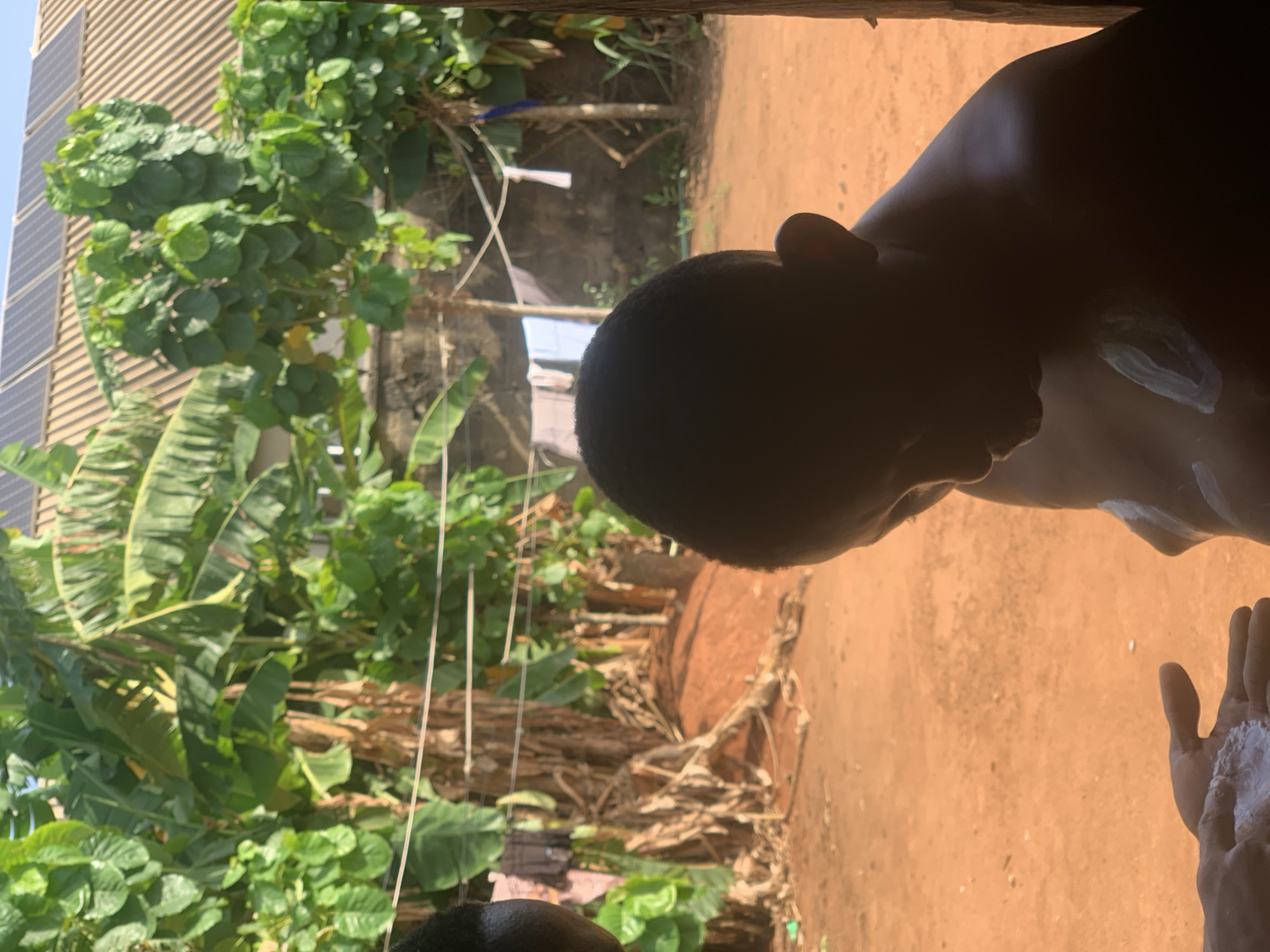 A person is seen from inside, looking outward at a lush, overgrown backyard. The view includes verdant foliage, banana trees, and a partial view of a building facade featuring solar panels. In the foreground, the person has a substance, possibly sunscreen or lotion, applied to their shoulder and held in their hand. The backyard transitions from the dense greenery to a more open, earthen area. The location is identified as Ikorodu, Nigeria.