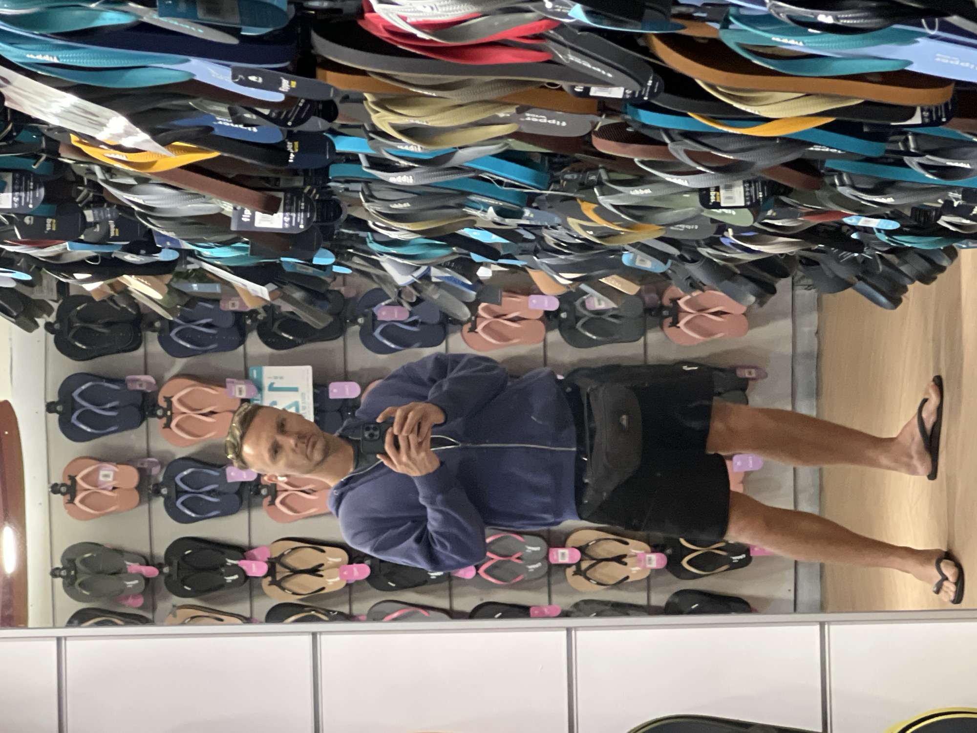 A man is standing in front of a display of flip-flops in what appears to be a store. The display has rows of flip-flops in various colors and styles, hanging on hooks. The man is wearing a blue zip-up jacket and black shorts. He is holding a phone and appears to be taking a picture or video. The reflection in the mirror suggests the store is located in Denpasar, Indonesia.