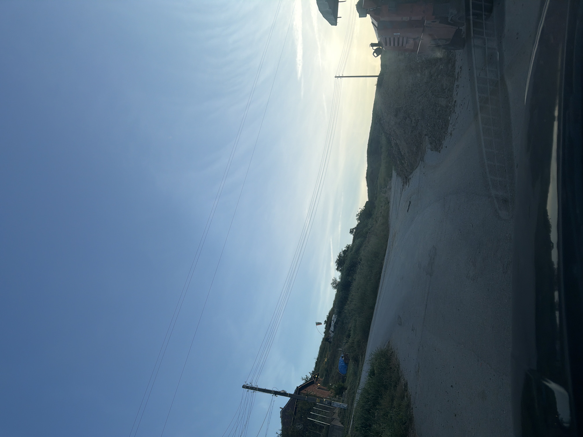 The image shows a view from a vehicle on a road near the city of Iskar, Bulgaria. To the right is a partially obscured road and a grassy embankment. A tractor is partially visible. Powerlines are running across the sky. In the distance, there is a building. The sky is blue and lightly cloudy.