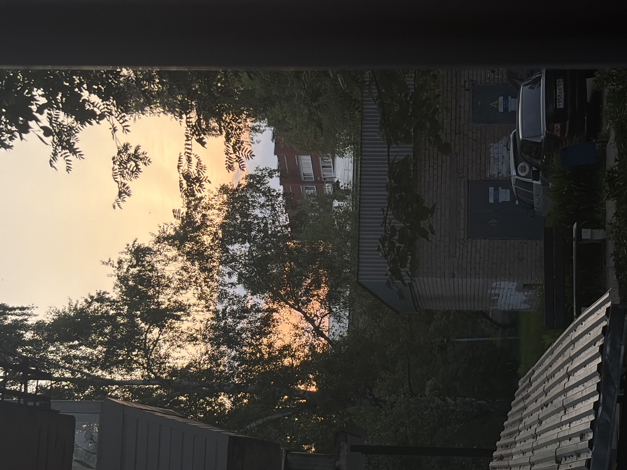 View through a window of a sunset sky partially obscured by trees and buildings. A multi-story brick building with a red roof is visible in the background. To the right, a brick building with garage doors is present, and a black car is parked next to it with license plate "Y958AA 152". In the foreground, parts of a building with corrugated roofing are visible. The location is an unknown urban environment.