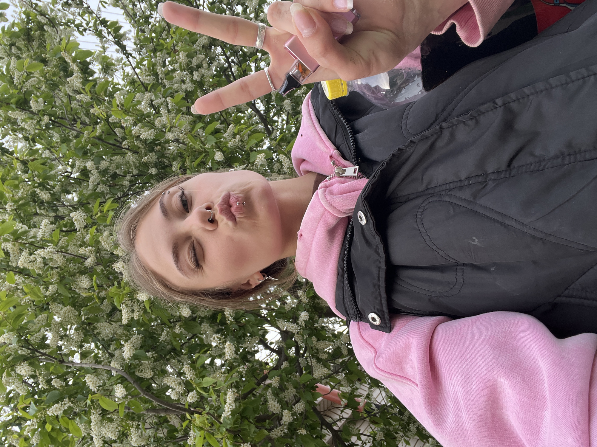 A woman poses in front of a flowering tree. She is fair-skinned with light brown hair. She wears a light pink hoodie under a black vest, with metal rings on her fingers. She has body modifications, including a nose ring, a lip piercing, and an eyebrow piercing. She makes a kissing face and holds up a peace sign. In the background is a tree in full bloom, with white flowers. Based on the visual elements there is nothing to suggest this image was taken in the city of Фоки, Russia.
