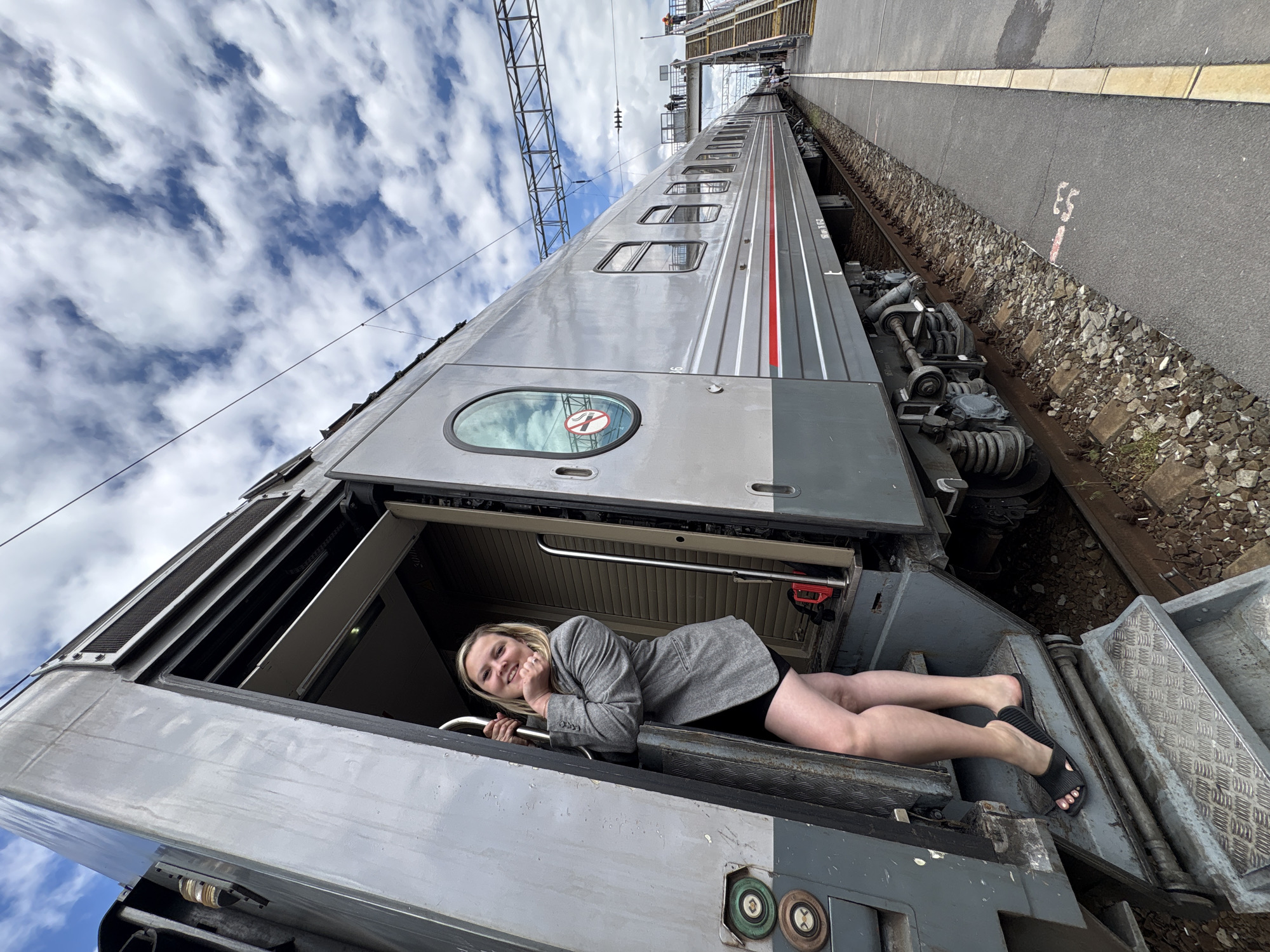 A wide-angle, high-angle shot depicts a woman lying in the open doorway of a train car at what is possibly a railway station. The train car is primarily silver and gray, with red accent stripes. The woman, who appears to be Caucasian, is wearing a gray blazer, a black skirt, and black sandals. She is propped up on her elbow, holding onto a handrail, and smiling towards the camera. The train is parked alongside a gray concrete platform with a gravel area and train tracks visible on the other side. Overhead power lines and structures are seen above the train. The sky is filled with puffy white clouds and patches of blue. Writing is on the pavement in red. The location is not able to be confirmed.