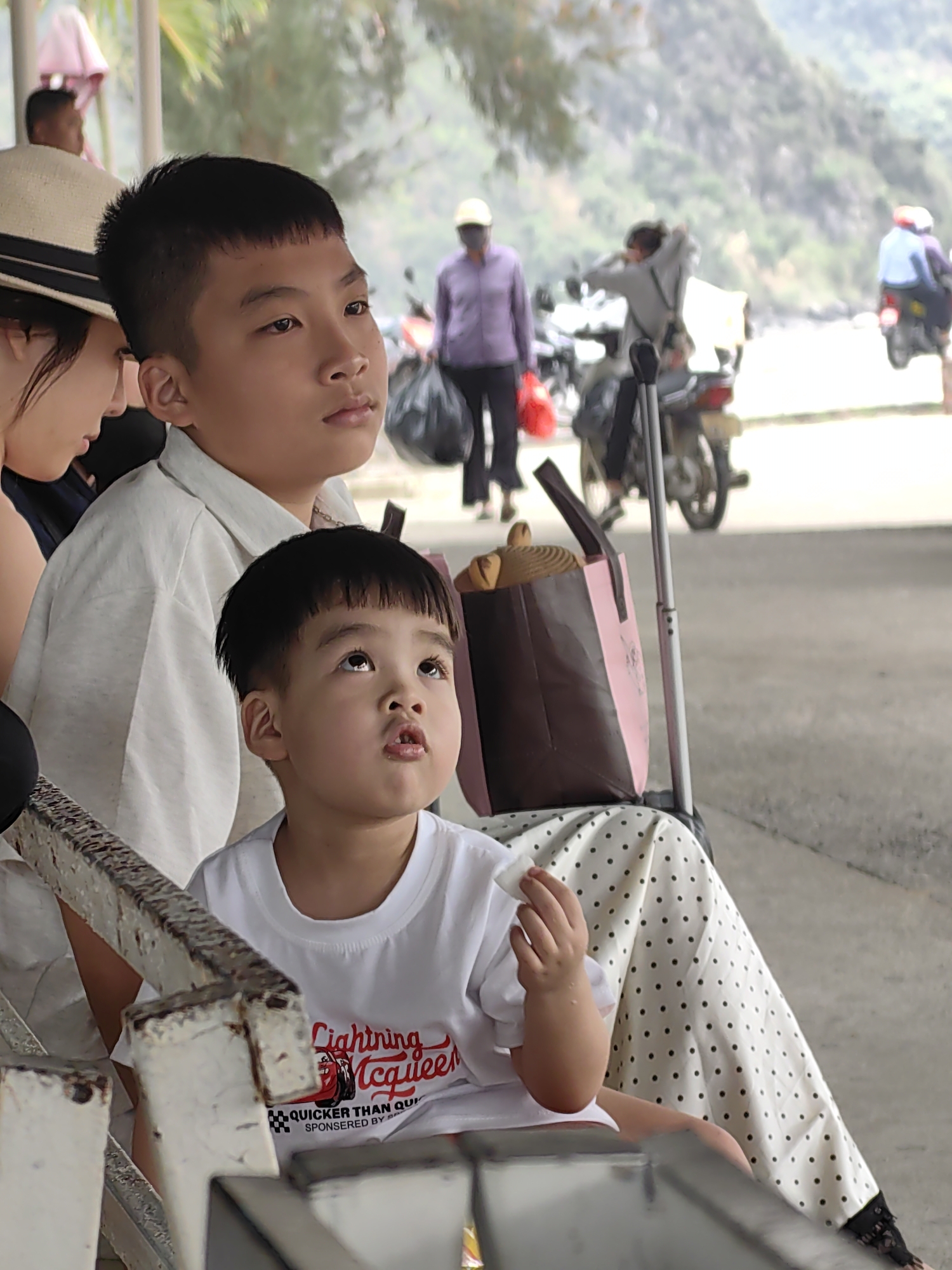 Three individuals are the primary focus: a woman wearing a hat and dark top, a pre-teen boy in a light-colored shirt, and a younger boy in a white "Lightning McQueen" t-shirt. All three are seated. The younger boy looks upwards, and the older boy gazes into the distance. Behind them is a bag containing baked goods, possibly croissants.
The background includes several people, including a woman in a purple shirt and figures on motorbikes. The scene is outside, adjacent to a road with mountains visible in the background. The location is likely Hải Phòng, Vietnam.
