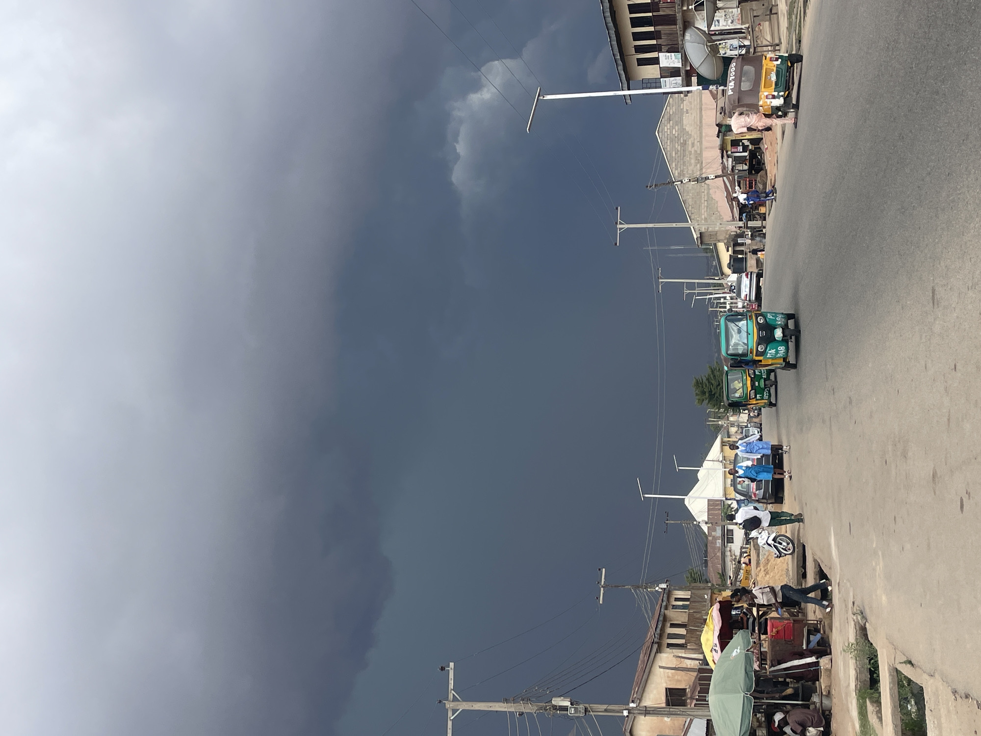The image depicts a street scene in Jos, Nigeria. A dark, ominous sky looms overhead. Along the right side of the image, a paved road stretches into the distance, with a variety of vehicles, including several auto rickshaws (tuk-tuks) in green, blue, and yellow. A few pedestrians are walking on the road. Adjacent to the road are buildings of varying heights and styles, some with visible rooftops and satellite dishes. Utility poles line the street, with wires stretching across the sky. A roadside vendor with an umbrella is visible in the foreground.