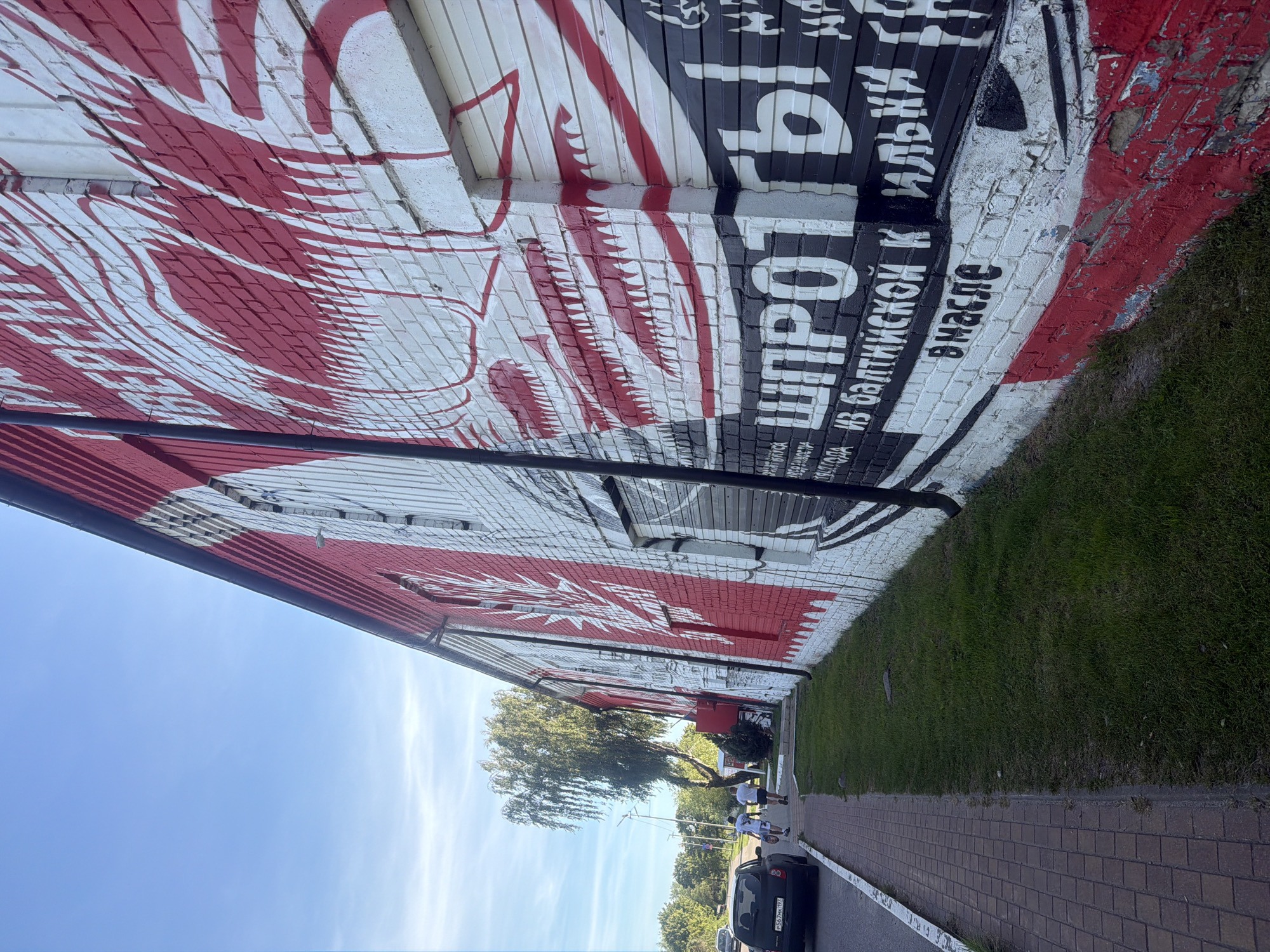 The image depicts a building with a large mural painted on its exterior brick wall, located in Vzmorye, Russia. The mural features red and white graphic elements, possibly depicting stylized fish or waves, and large white Cyrillic text. Below these elements, there is black text on a white background that reads "ШПРОТЫ ИЗ БАЛТИЙСКОЙ К ИЛЬКИ В МАСЛЕ", which translates to "Sprats from Baltic Kilka in Oil." The lower portion of the mural shows red paint peeling off the brick. To the left of the building, a sidewalk made of brick pavers runs alongside a strip of green grass. A black car is parked on the street next to the sidewalk. Two individuals, appearing to be adult males wearing t-shirts and shorts, are walking away from the camera on the sidewalk. A street light and a deciduous tree are visible behind them. The sky is blue with scattered clouds.