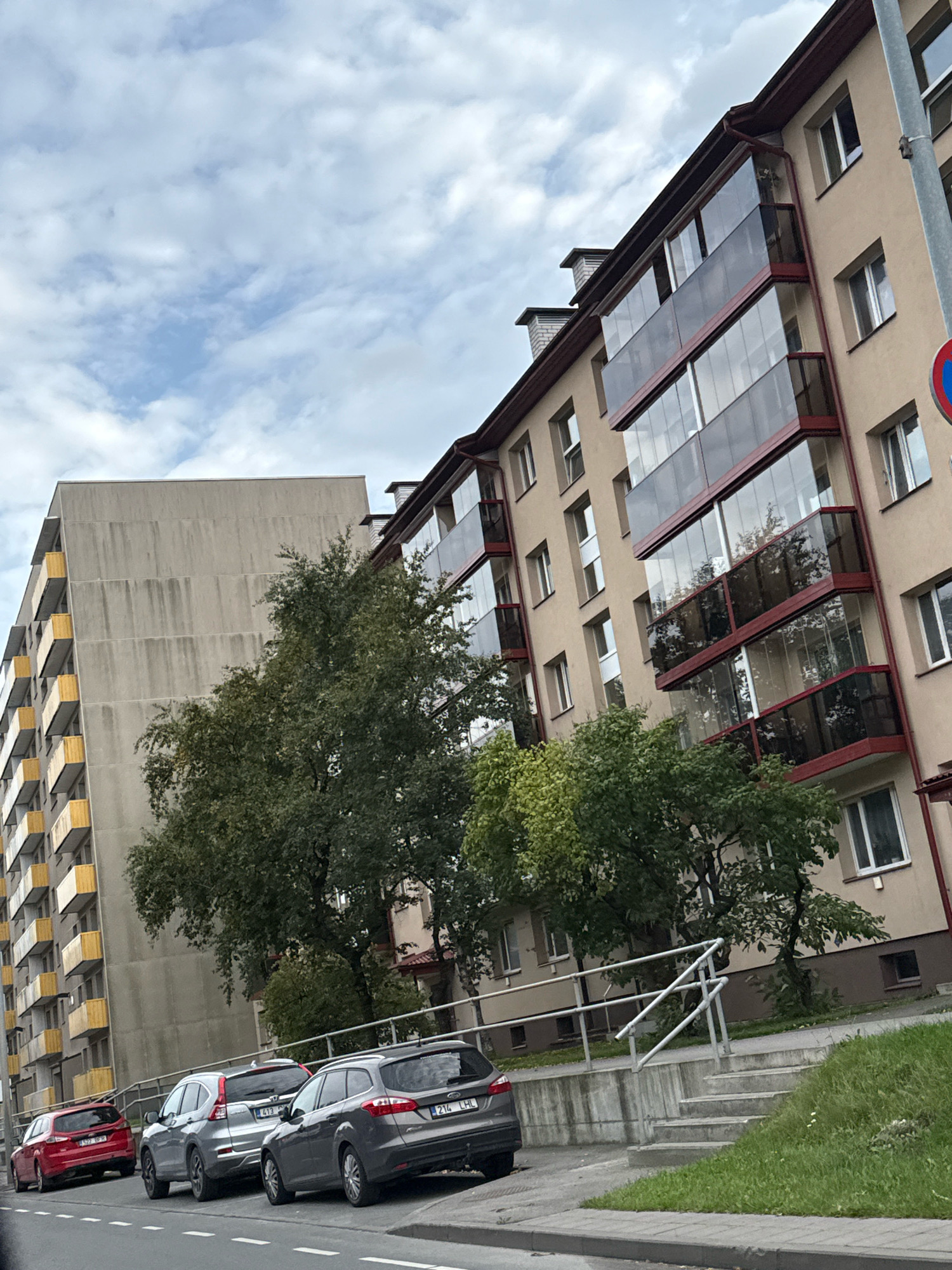 The image shows a residential building in Tallinn, Estonia. The building has a beige facade and features multiple balconies with glass enclosures. In front of the building, there is a row of parked cars, including a red hatchback, a silver SUV, and two gray station wagons. A concrete staircase with a metal railing leads up from the sidewalk. Trees and greenery are visible around the building. The sky is cloudy.