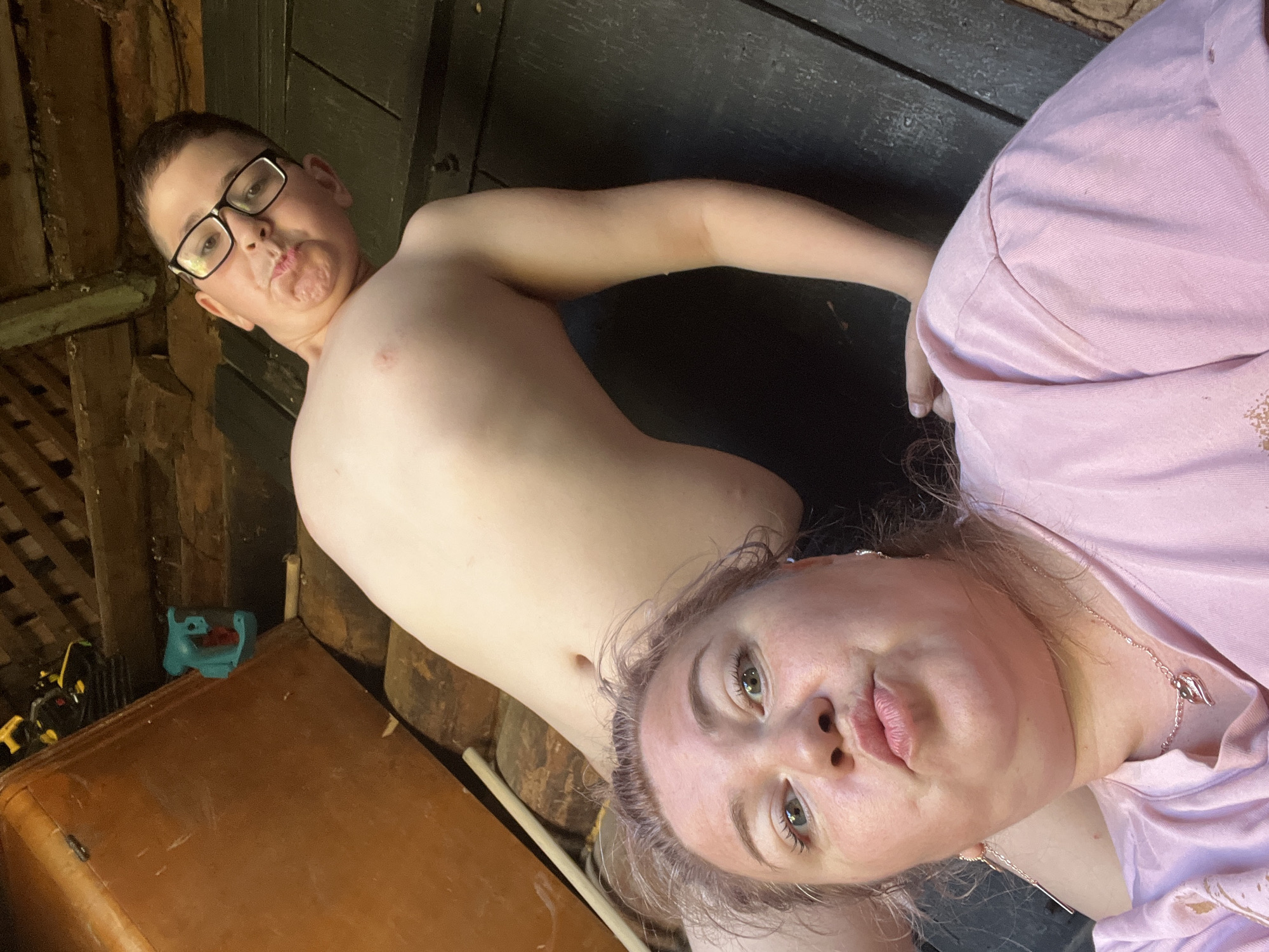 The image depicts a selfie taken by a woman in the foreground, with a young boy in the background. The woman has light-colored hair and is wearing a pink t-shirt. She is making a kissing face. The boy, wearing glasses, is positioned behind her and also appears to be making a funny face. They are in an indoor environment with wooden structures and a large, reddish-brown object that may be furniture. The floor appears to be made of dark, weathered planks. There is a teal-colored toy on top of the reddish-brown object. The overall impression is that they are in a rustic setting, possibly a shed or an unfinished room. The location is identified as Фоки, Russia.