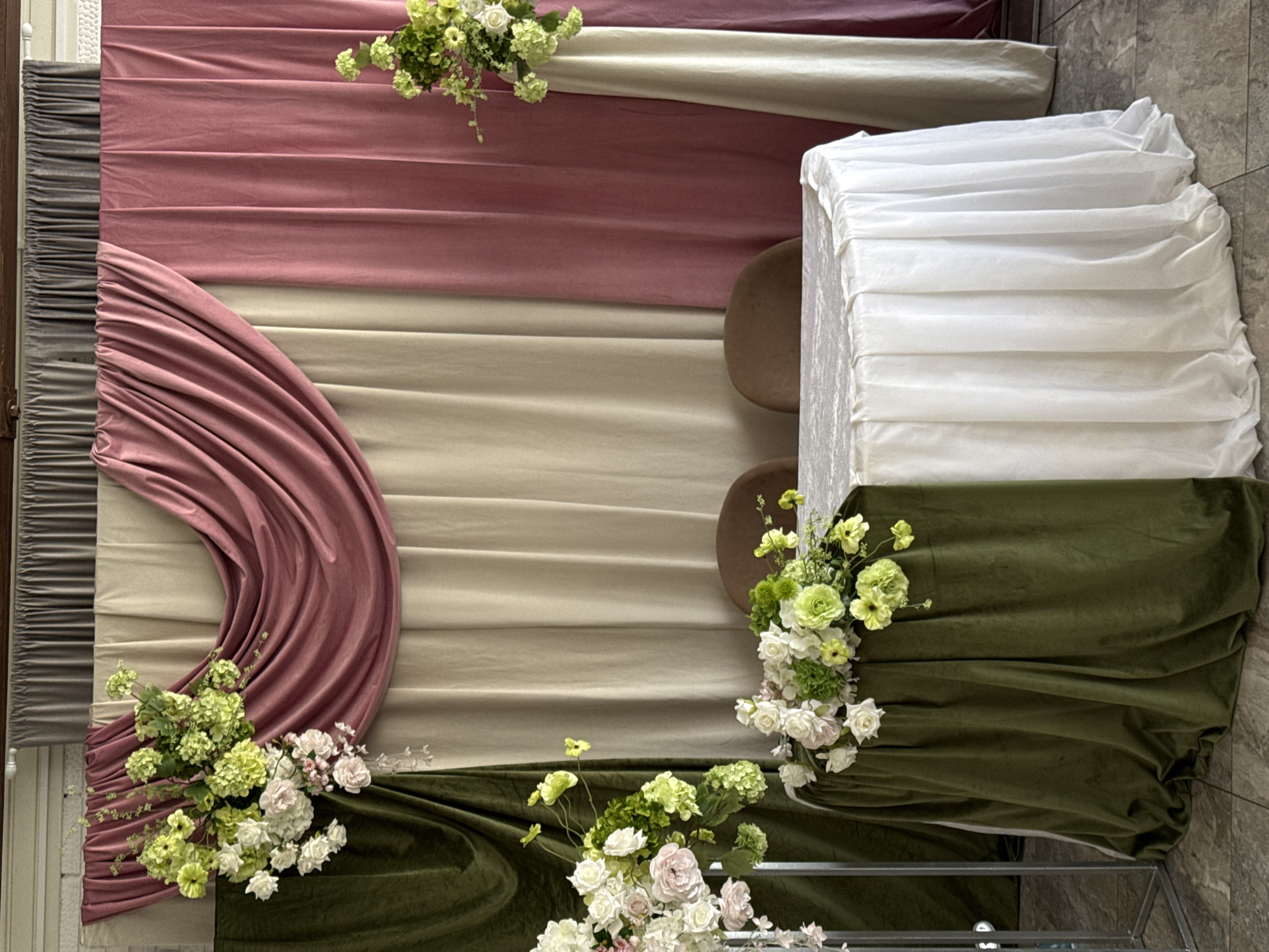 The image shows a decorated table set for two, likely for a special occasion, in what appears to be an indoor venue. The backdrop is composed of layered drapes in varying shades of pink, beige, and dark green. A large floral arrangement with white and green flowers is prominently displayed to the left of the table. The round table is covered with a white tablecloth that drapes down to the floor, with a partial green drape on the left side. Two chairs are visible behind the table. The flooring appears to be tiled. There are no people present in the image. There is no information in the image that indicates this scene is located in Votkinsk, Russia.