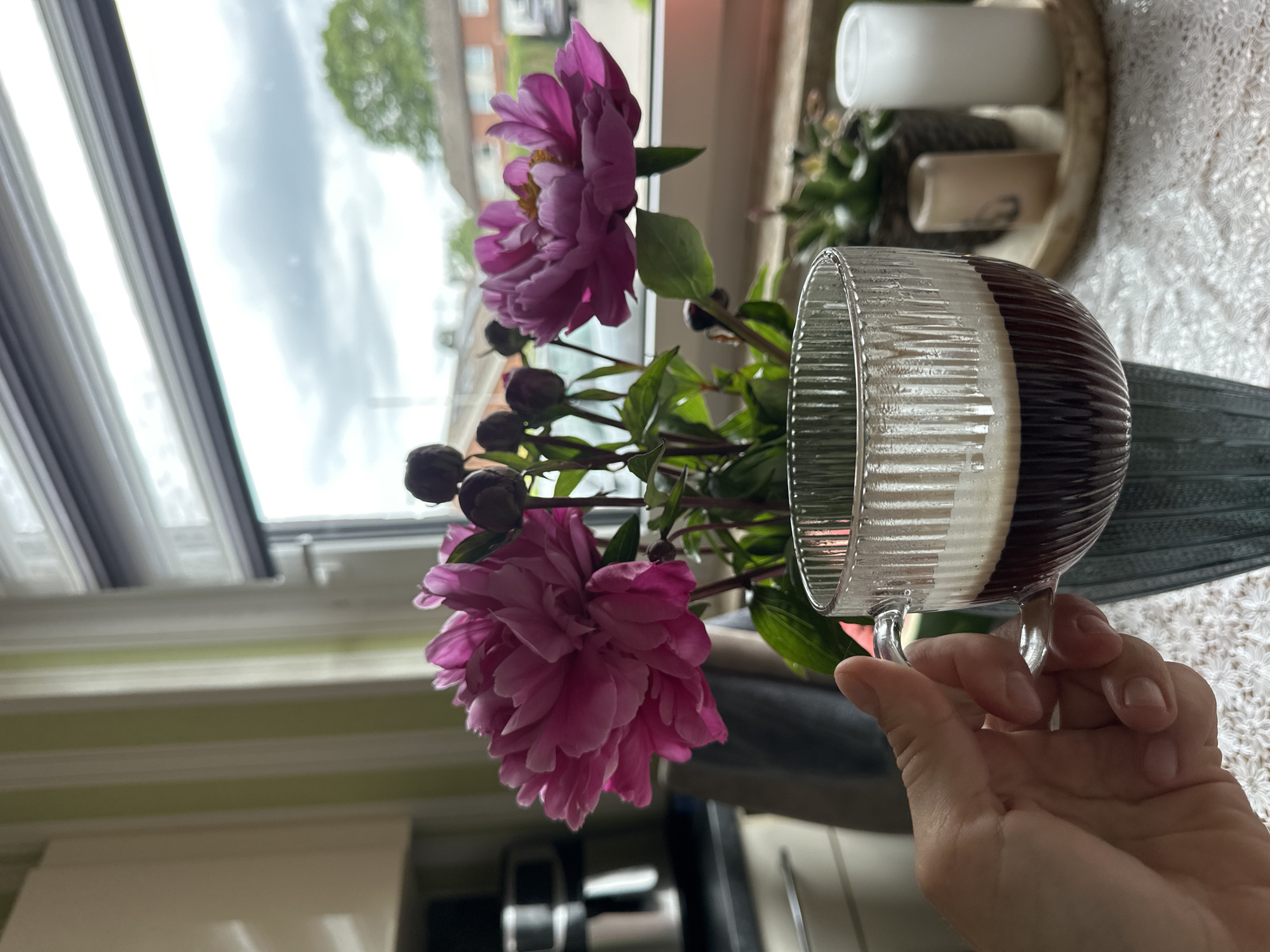 A close-up, eye-level shot shows a person's hand holding a glass mug of coffee. The mug has vertical ridges and is filled with dark liquid topped with foam. The mug sits on a table with a white lace tablecloth. Behind the mug, a bouquet of vibrant pink peonies and their buds are displayed in a textured blue vase. To the right of the flowers, a small potted plant and two white cylindrical objects, possibly candles, are arranged on a light-colored tray. The background is a window with horizontal blinds. Through the window, a cloudy sky and a residential area with houses and trees are visible, suggesting the scene takes place in Fredenbeck, Germany. The lighting appears to be natural, coming from the window, creating shadows on the left side of the image. The overall impression is a still life composition with elements of nature and everyday life.