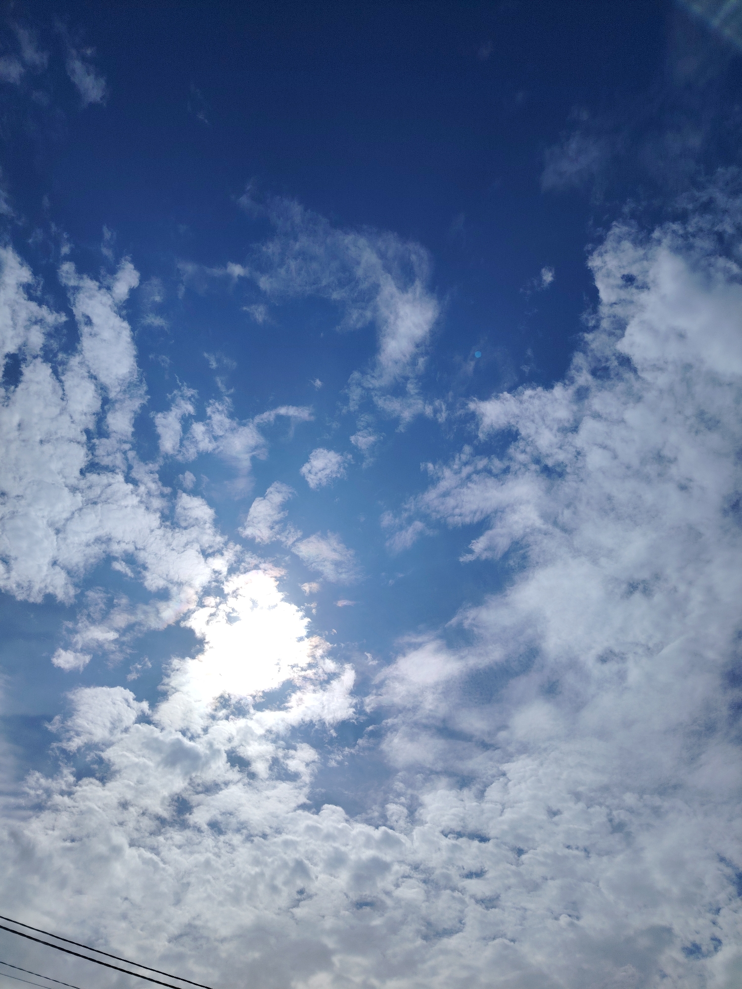 The image depicts a bright, sunny sky with scattered white clouds, taken from Severynivka, Ukraine. The sun is partially obscured by clouds, creating a radiant glow. The bottom of the frame shows a portion of two dark power lines, suggesting an outdoor, possibly rural or suburban, setting. The sky is a deep blue, with varying densities of clouds, some wispy and translucent, others more solid and opaque. The overall impression is one of a clear, pleasant day.