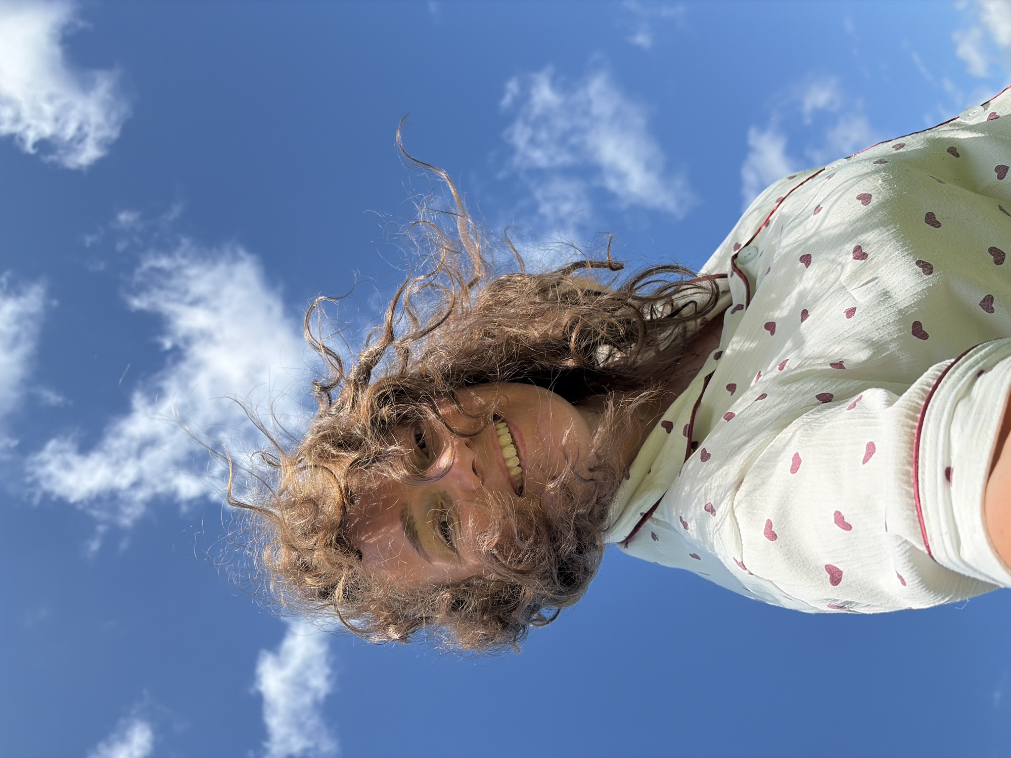 A low-angle, medium shot captures a young woman with curly brown hair smiling directly at the camera. Her hair is windswept, framing her face and obscuring parts of her left side. She is wearing a light-colored pajama top with a pattern of small, dark pink hearts and a burgundy trim along the collar and placket. The background is a vibrant blue sky with scattered white clouds. The image appears to be taken outdoors on a sunny day, likely in Ольховка, Russia, given the context. The woman's expression is cheerful and relaxed.