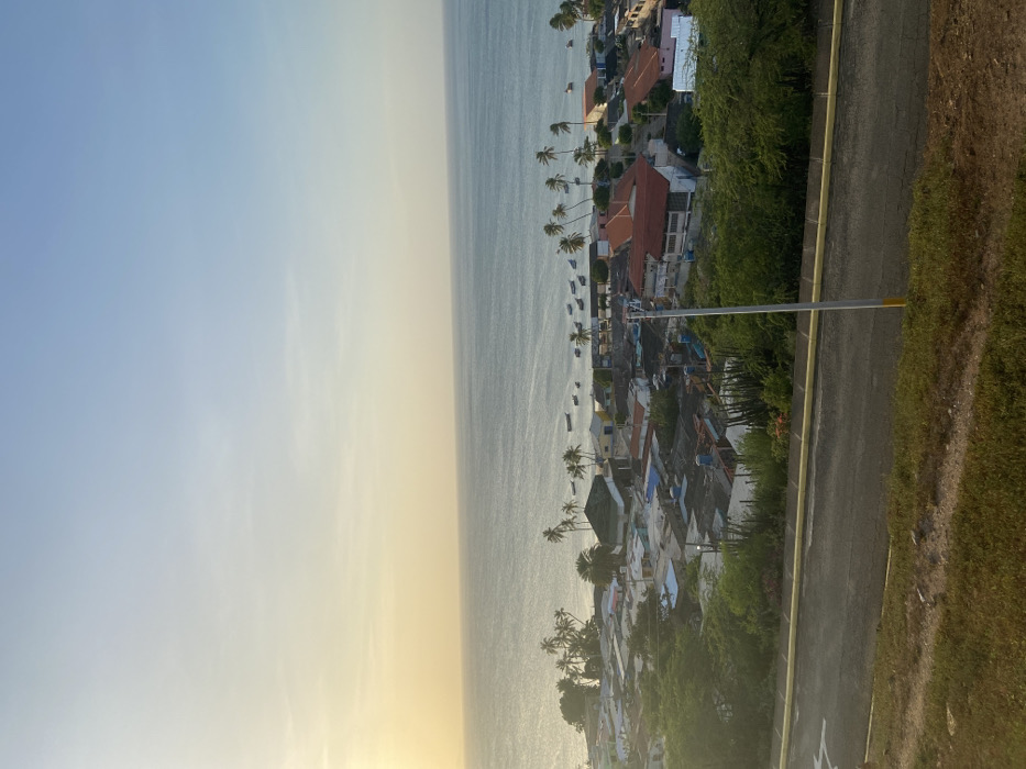 A coastal town is captured from an elevated perspective with the ocean on the left and a road on the right. Buildings, many with red roofs, are nestled among palm trees. Boats dot the water near the shoreline. The sky fades from blue to a warm, golden hue near the horizon. The road is flanked by green vegetation, transitioning into dry grass on the outer edge. A lamppost stands beside the road, extending into the frame.