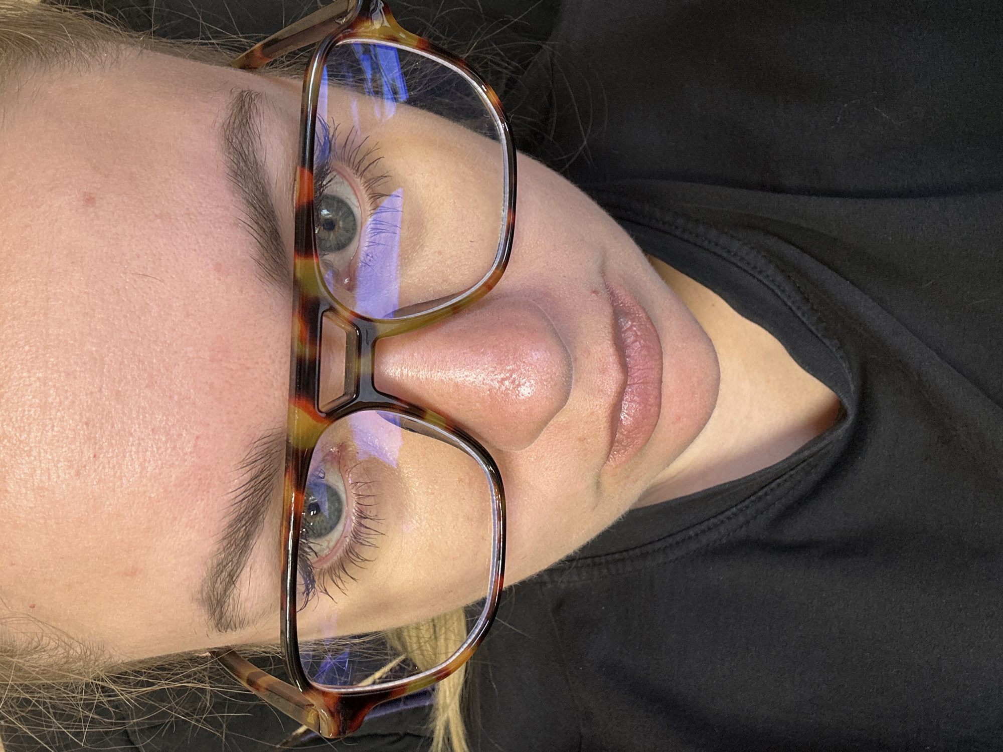 A close-up, angled portrait of a woman wearing eyeglasses. The eyeglasses have a tortoiseshell frame. The woman has fair skin, light eyebrows, and blue eyes with visible eyelashes. Her lips are closed. She is wearing a black top. The background is indistinct. The image provides no information to indicate it was taken in Ural'skoe, Russia.