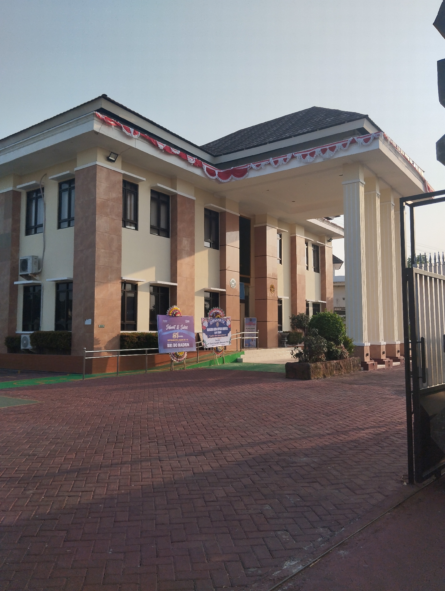 The image depicts the exterior of a two-story building, likely a government or institutional office, in Madiun, Indonesia. The building features a beige and brown facade, with prominent white pillars supporting a covered entryway. Red and white festive banners are draped along the eaves of the roof. In front of the building, there is a paved walkway made of red brick pavers, and several potted plants and shrubs are visible. Two signs are placed near the entrance, one purple and one blue, with decorative floral arrangements. A black metal gate is partially visible on the right side of the frame. The sky is clear and blue, suggesting daytime. The overall scene conveys a formal and well-maintained environment.