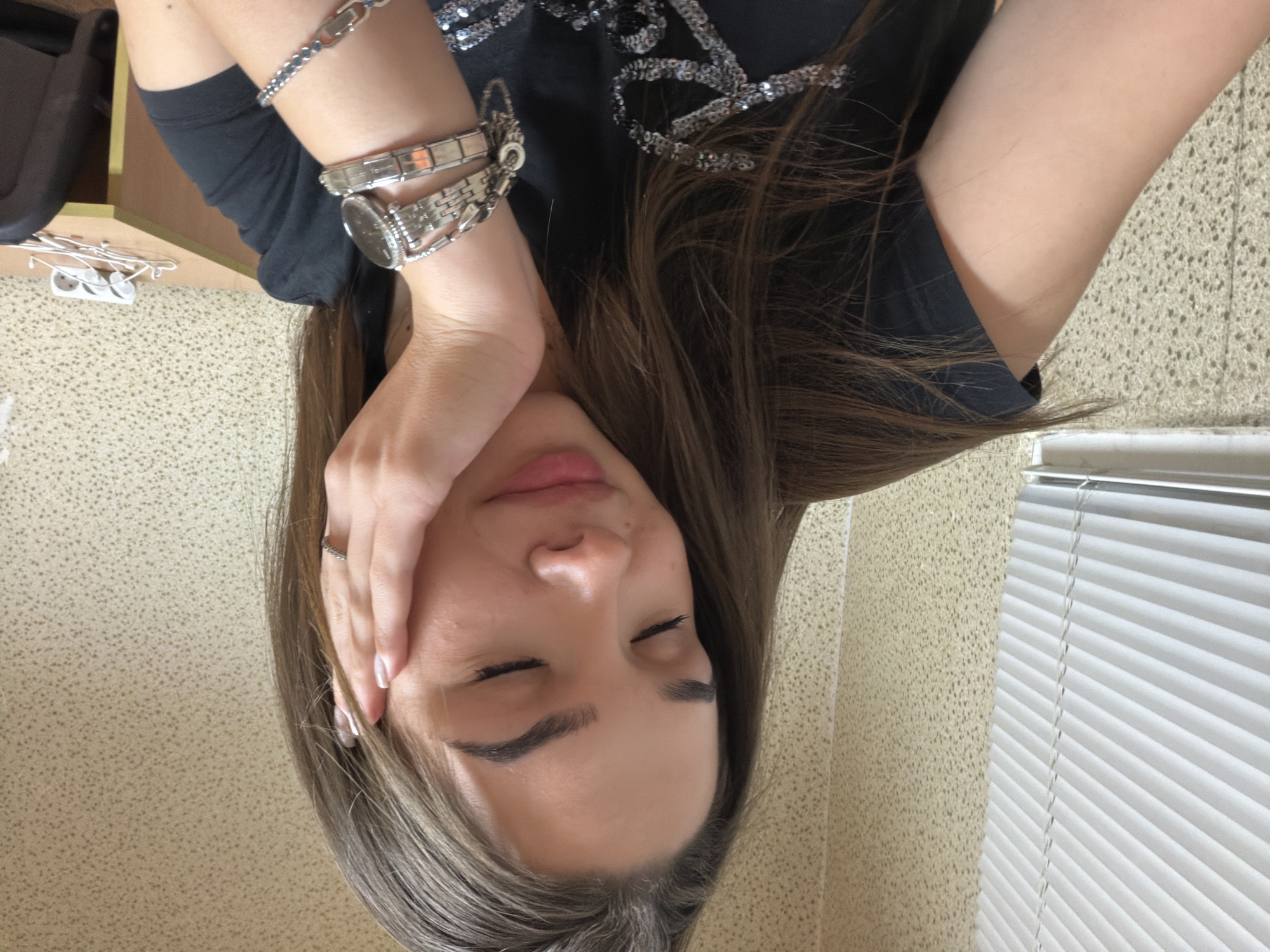 A woman is seen from an upside-down perspective, eyes closed, with a hand resting on her face. She wears a silver watch and bracelets. Her hair is light brown. She is wearing a black top with a sequin design. The background includes a textured wall and a window with white blinds. The location is not discernible from the image alone.
