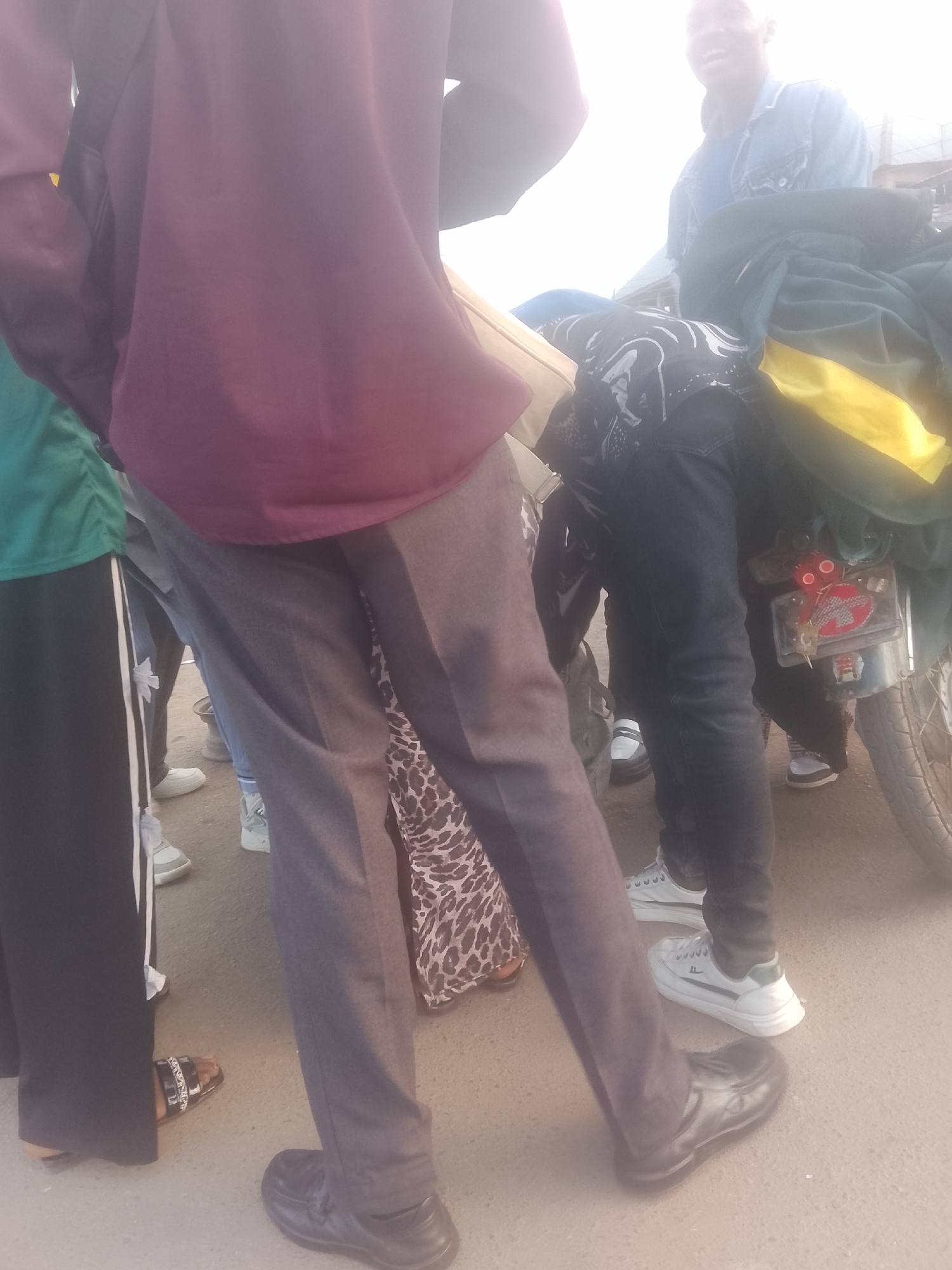 The image depicts a group of people and a motorcycle in what appears to be an outdoor setting in Gwong, Nigeria. Several individuals are partially visible, wearing various clothing items. One person is wearing a maroon shirt and gray pants with black shoes. Another person is wearing a green shirt with black pants and sandals. A third person is wearing a jean jacket and is sitting on the motorcycle. The motorcycle is loaded with items covered in a green and yellow fabric. The setting is a street or similar paved area.