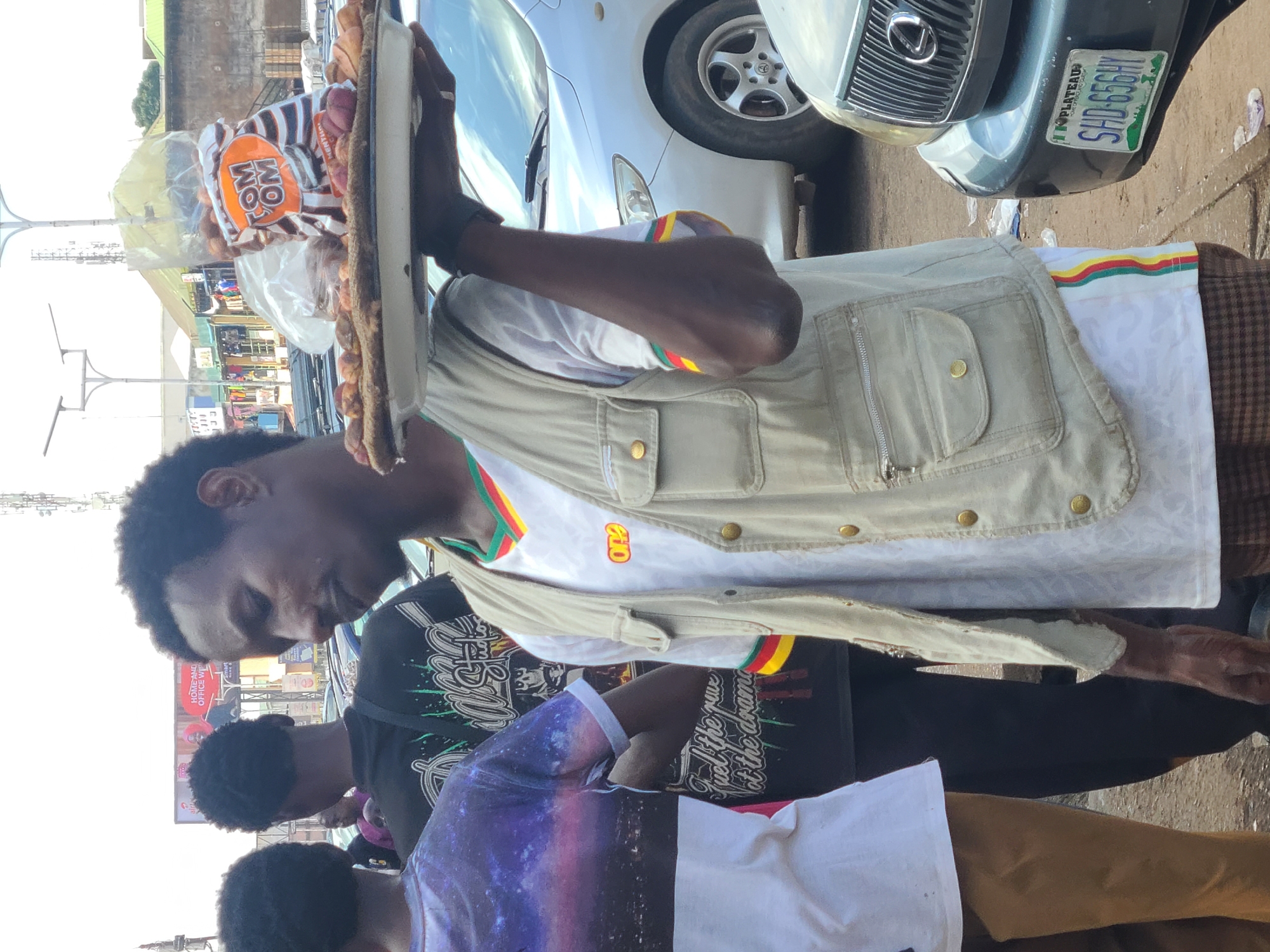 This media file captures a vibrant street scene in Jos, Nigeria, focusing on a young man balancing a tray of pastries on his head. He is dressed in a light-colored vest over a white shirt with a colorful trim, and he appears to be moving through a busy outdoor area. Behind him, several other individuals are visible, some also carrying items.
The background reveals a street environment with vehicles, including a car with a visible license plate that reads "SHD-656HY PLATEAU". This suggests the location is likely Nigeria, and specifically Plateau State. There are also shops or market stalls in the distance, indicating a commercial area. The overall impression is that of a typical day in a bustling Nigerian city.
The pastries on the tray are mostly round, baked goods, with some packaged items labeled "TOM OM". The lighting suggests it's daytime, possibly mid-morning or afternoon, with the sun casting some shadows. The individuals in the scene appear to be engaged in their daily activities, with no overt display of strong emotions. The image conveys a sense of everyday life and commerce on a Nigerian street.