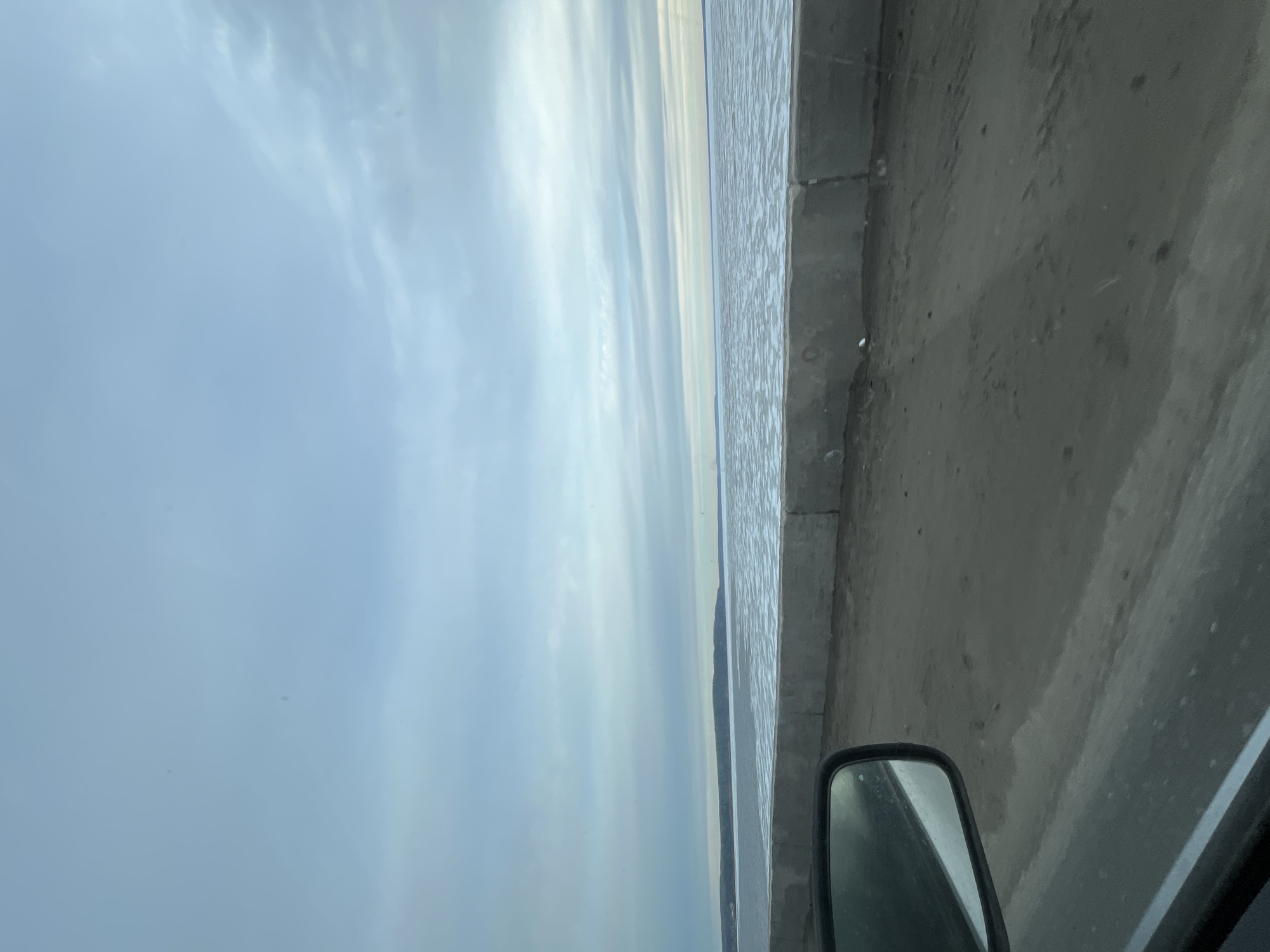 The image appears to be taken from inside a vehicle, possibly a car, driving along a road next to a body of water. The sky is overcast with light clouds, and the water appears calm. A concrete barrier separates the road from the water's edge. A side mirror of the vehicle is visible in the lower right corner. There is no information about the location of the image being from the city of Chaikovsky, Russia.