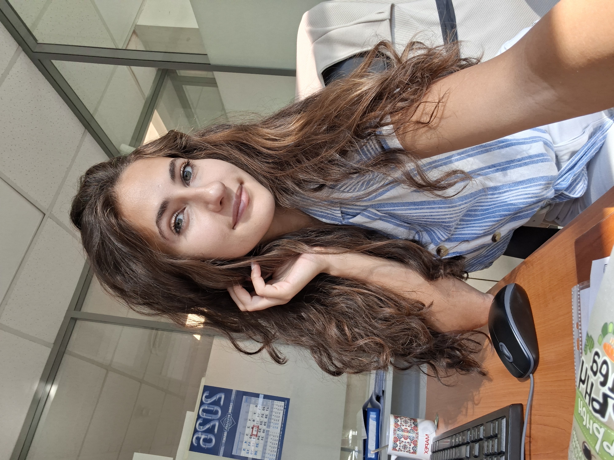 A young woman with long, wavy brown hair and blue eyes is captured in a selfie taken from a slightly high angle. She is seated at a wooden desk in what appears to be an office setting. Her hair is partially covering her left side, with strands framing her face and some falling over her shoulder. Her right hand is raised, with fingers gently touching her hair near her ear. She is wearing a blue and white striped shirt.
On the desk, a black computer keyboard and a matching computer mouse are visible to the left. A white mug with a colorful, patterned design sits to the left of the keyboard. Behind the keyboard and mug, a blue folder and a calendar with the ASOS logo are affixed to the wall. The background consists of a modern office environment with glass partitions, a ceiling with acoustic tiles, and a portion of an office chair visible on the right. Sunlight streams in from the right, casting a shadow and highlighting the textures in the scene. The overall impression is one of a casual moment captured in a professional workspace.