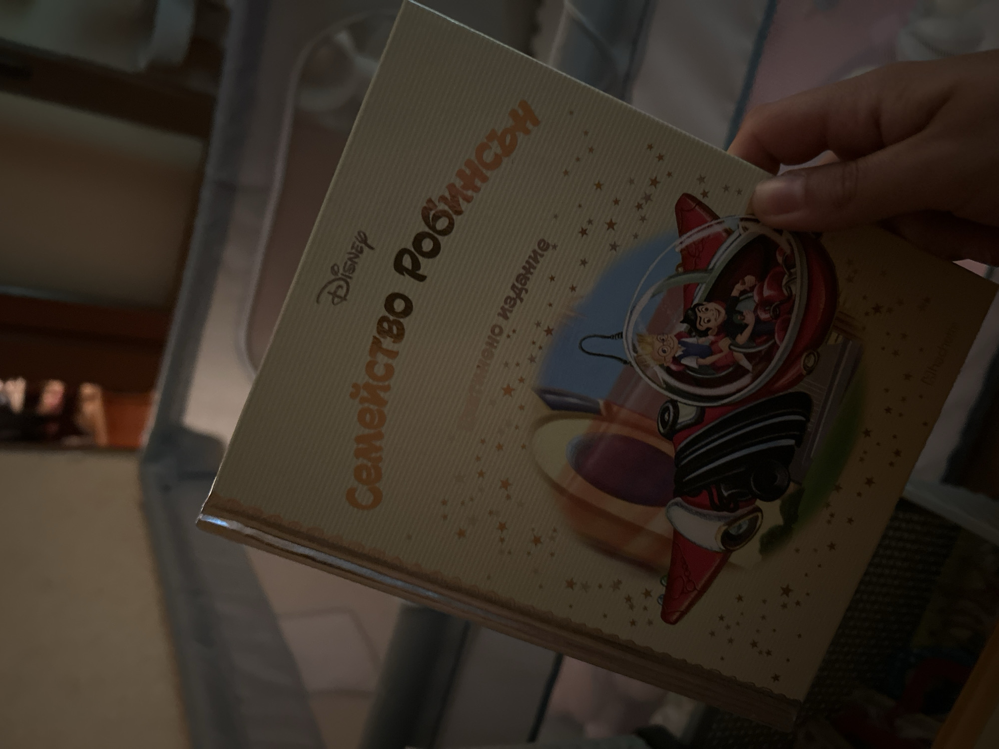 The image shows a book titled "Семейство Робинсън" (Family Robinson) by Disney. The book cover features an illustration of two boys in a red vehicle, possibly a rocket or airplane. The book is being held by a person's hand, with the index finger and thumb visible. The background appears to be a dimly lit room, possibly a nursery or bedroom, with a crib or playpen visible. There are no visible landmarks or clues to suggest a specific location within Sofia, Bulgaria. The scene is indoors, with artificial lighting.