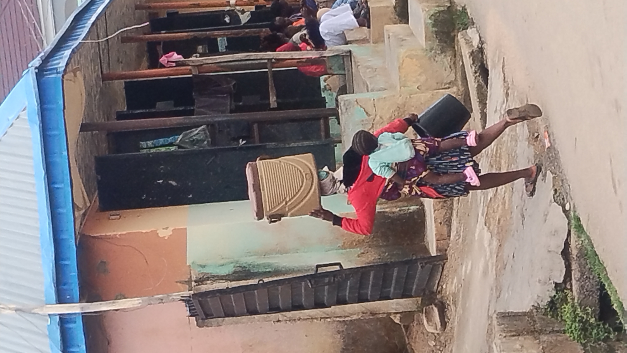 The image depicts an outdoor scene featuring a woman walking on a stepped pathway. The woman, dressed in a red long-sleeved top and a patterned skirt or wrapper, carries a child on her back secured by fabric. In her right hand, she holds a black bucket, and in her left, a light tan or beige plastic object, possibly a portable stool or a laundry basket. She is wearing sandals.
The pathway consists of concrete steps and surfaces, leading upwards. In the background, multiple levels of structures are visible, with more steps and platforms where several other people are present, some appearing to be sitting or resting. One individual in the background is wearing white clothing. The surrounding architecture suggests a modest, possibly residential or communal setting, characterized by plain walls and simple construction. To the far left, a structure with blue-painted trim and corrugated metal roofing is partially visible. The scene is well-lit, indicating it is daytime. No visible text is present in the image.