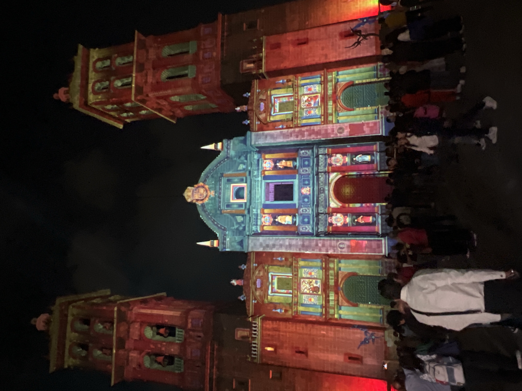 A low-angle, long shot shows a large group of people milling around two buildings with projection-mapped exteriors on a night in Unknown. The buildings stand close together, with the taller one in the center of the shot. Both are brick-colored, with two tall, thin towers to each side. The projection mapping covers the buildings in intricate designs that resemble stained glass windows. The colors of the designs are primarily red, green, and blue. The people are of various ages and ethnicities. They are dressed in casual clothing and appear to be enjoying the light show. The overall tone of the image is festive and energetic.