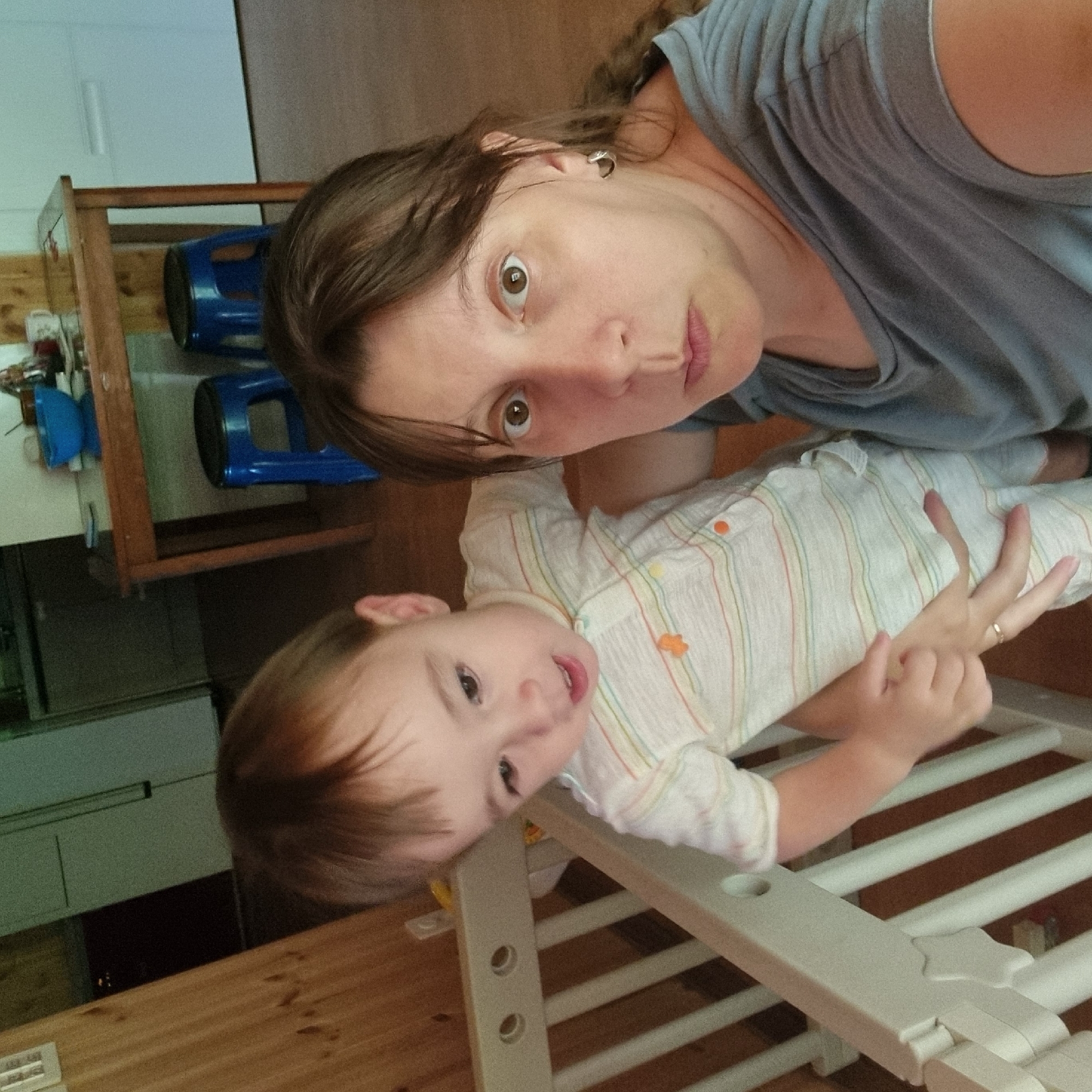 In this indoor photograph, a woman and a young child are captured from the chest up. The woman, on the right, faces the camera with wide, startled eyes. She has brown hair and is wearing a grey t-shirt. The child, to the left, is dressed in a light-colored onesie with thin horizontal stripes. The child's body is partially obscured by the woman's arm, and the child appears to be reaching out. A portion of a light-colored crib or playpen is visible in the foreground. The background shows a wooden wall and furniture, including a wooden table with blue stools and a blue bowl, suggesting a domestic setting. The image is taken from a slightly low angle. The scene is not identifiable as being in Dunnae-myeon, South Korea.