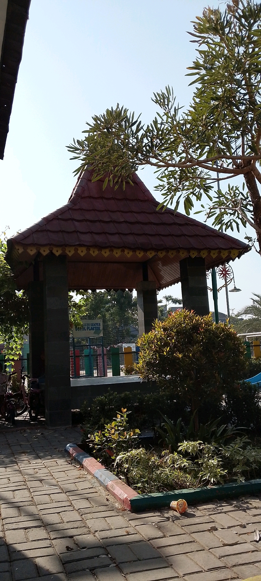 The image depicts an outdoor scene, likely in a park or public space in Satriamekar, Indonesia. In the foreground, a paved walkway made of grey rectangular bricks leads towards a structure. To the right of the walkway, a low border delineates a garden bed with various green plants. A single orange plastic cup lies on the paving stones near the garden bed.
The central focus of the image is a gazebo-like structure with a dark red, tiled, multi-tiered roof. The roof has decorative yellow trim. The gazebo is supported by four dark grey stone pillars. Through the open sides of the gazebo, a fence with colorful vertical posts (red, green, yellow, blue) is visible, along with a sign that reads "BUANG SAMPAH BOTOL PLASTIK" (Dispose of plastic bottle waste). Behind the fence, more greenery and possibly buildings can be seen.
To the left of the gazebo, a bicycle with red handlebars is parked on the brick walkway. Trees with green leaves are present on either side of the gazebo, with one tree extending over the roof. The sky above is clear and light blue. The overall impression is a well-maintained outdoor recreational area.