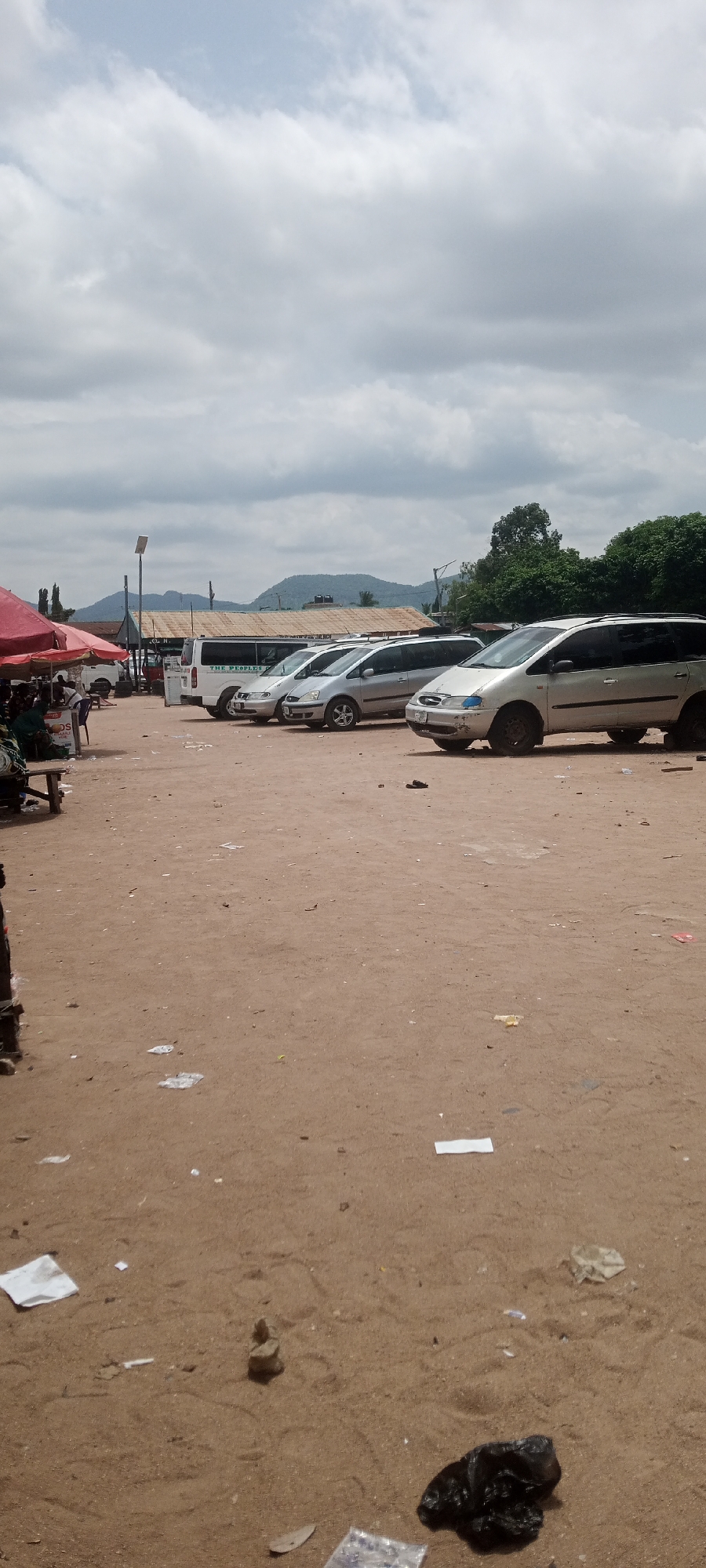 An outdoor, daytime, ground-level shot of a dusty, sand-covered area that appears to be a parking lot or a market hub. Several vehicles, including vans and SUVs, are parked in a row. In the foreground, there is debris scattered on the ground, including plastic bags, paper, and small stones. To the left, under a red canopy, a few people are visible sitting on a bench, with some goods displayed. In the background, there are buildings with tiled roofs and trees, and further in the distance, hills are visible under a cloudy sky. This scene is consistent with a transportation hub or market area within Jalingo, Nigeria, given the presence of multiple passenger vans commonly used for public transport in the region.