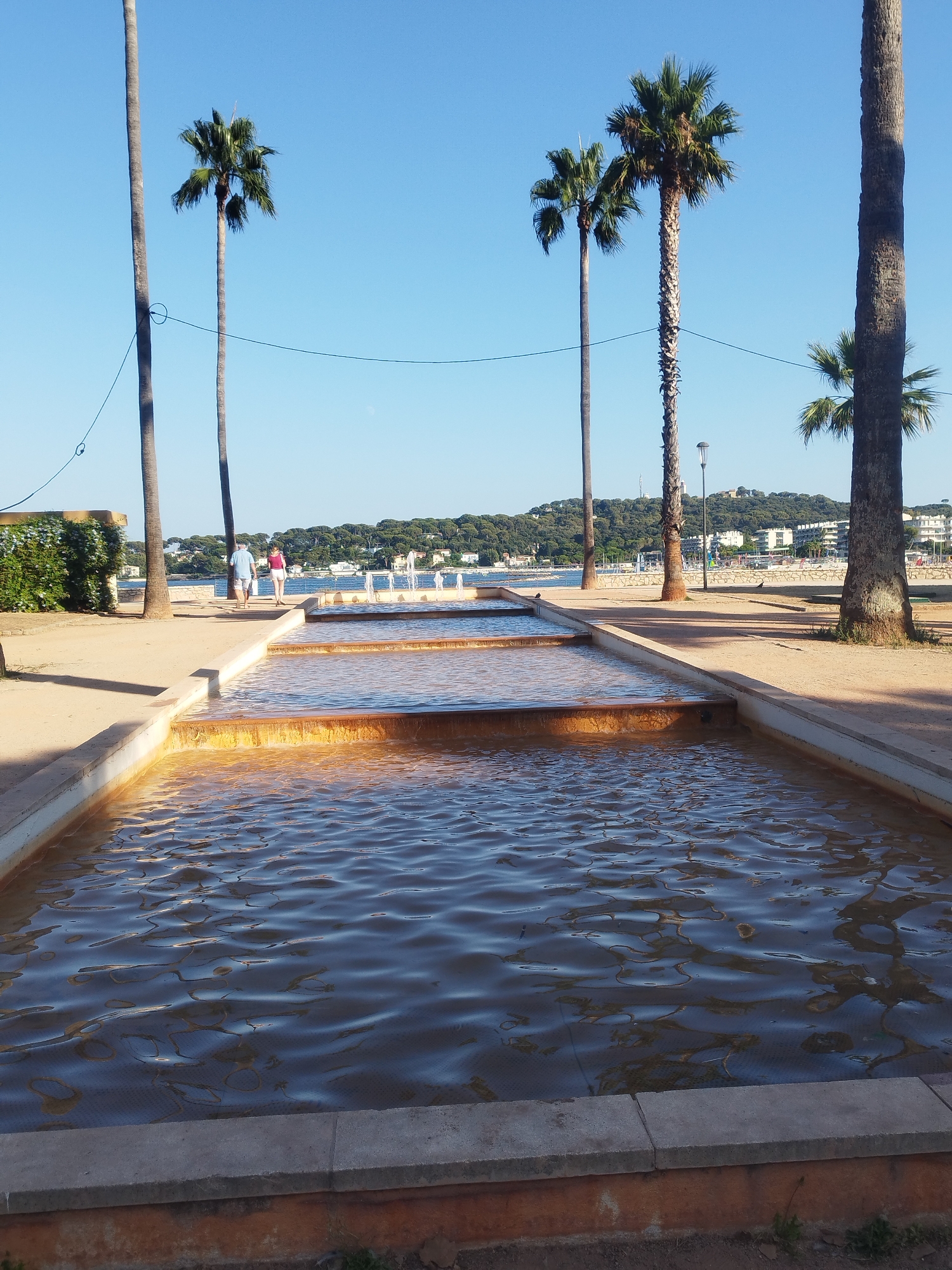 A tiered fountain with gently flowing water is the main subject in the foreground of this image. The water is a warm, golden color, reflecting the sunlight. Palm trees line a sandy pathway behind the fountain, with their fronds reaching towards a clear blue sky.
In the midground, two people are walking away from the camera on the pathway. The person on the left is wearing a blue shirt and shorts, and the person on the right is wearing a pink top and white shorts. Beyond them, a body of water, likely the sea, is visible, with a town or city nestled on a tree-covered hillside in the distance. Buildings and boats are scattered along the coastline. The overall scene suggests a pleasant, sunny day, likely in the late afternoon given the warm light. The location appears to be a seaside promenade or park area, characteristic of Antibes, France. The atmosphere is peaceful and relaxed, with the gentle movement of water and the presence of people enjoying the outdoors.