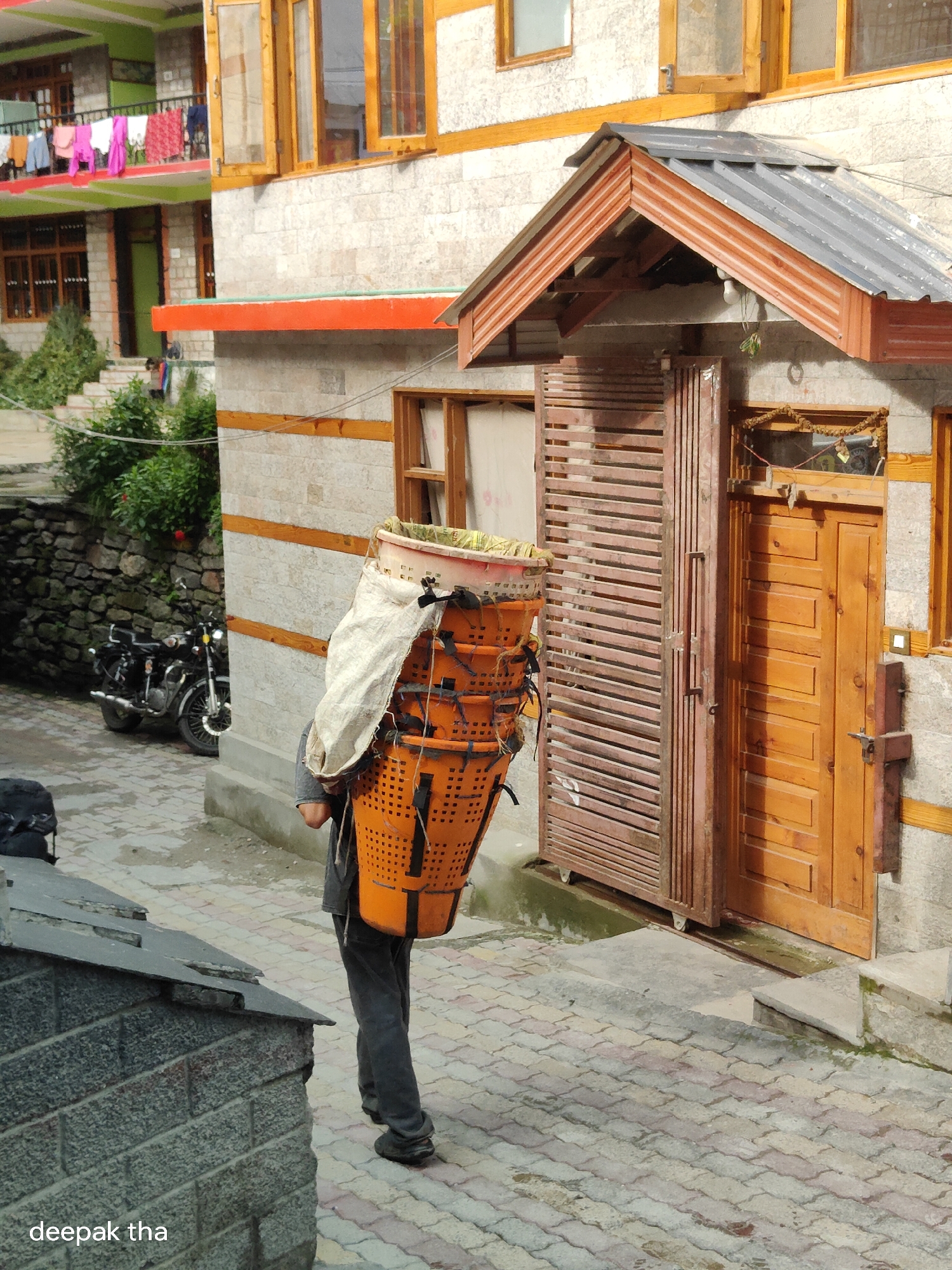 A person, likely a local, carries a large stack of orange plastic baskets woven with a white tarp over their shoulders in the narrow street of Vashisht, India. The baskets are tall and appear to be used for carrying goods. The person is walking away from the camera, wearing dark pants and dark shoes. Behind the person, a wooden gate and a brown wooden door are visible. To the left of the person, a black motorcycle is parked near a stone wall. The street is paved with interlocking bricks. The background features a building with traditional architecture, including wooden window frames and a red-orange trim. Clothes are drying on a balcony in the upper left portion of the image. The scene depicts a common sight in the village, highlighting local commerce and daily life.
