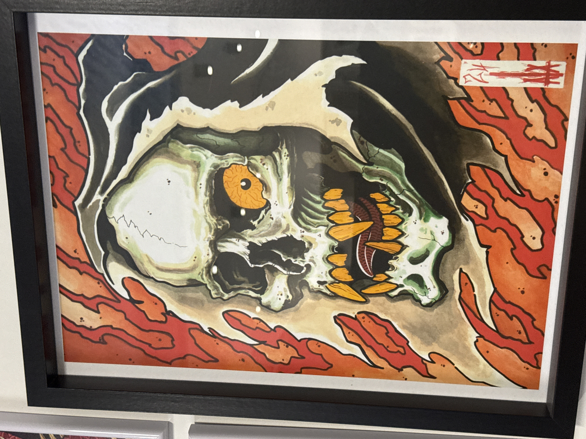 A framed artwork depicting a skull with an orange eye and pointed yellow teeth, framed by flames, is displayed on a wall in Sofia, Bulgaria. The artwork is positioned on a neutral-colored wall, with a black frame surrounding it. The overall aesthetic is that of a traditional tattoo or illustrative style. There are no people or specific identifiable locations in Sofia visible in the image. The focus is solely on the artwork itself.
