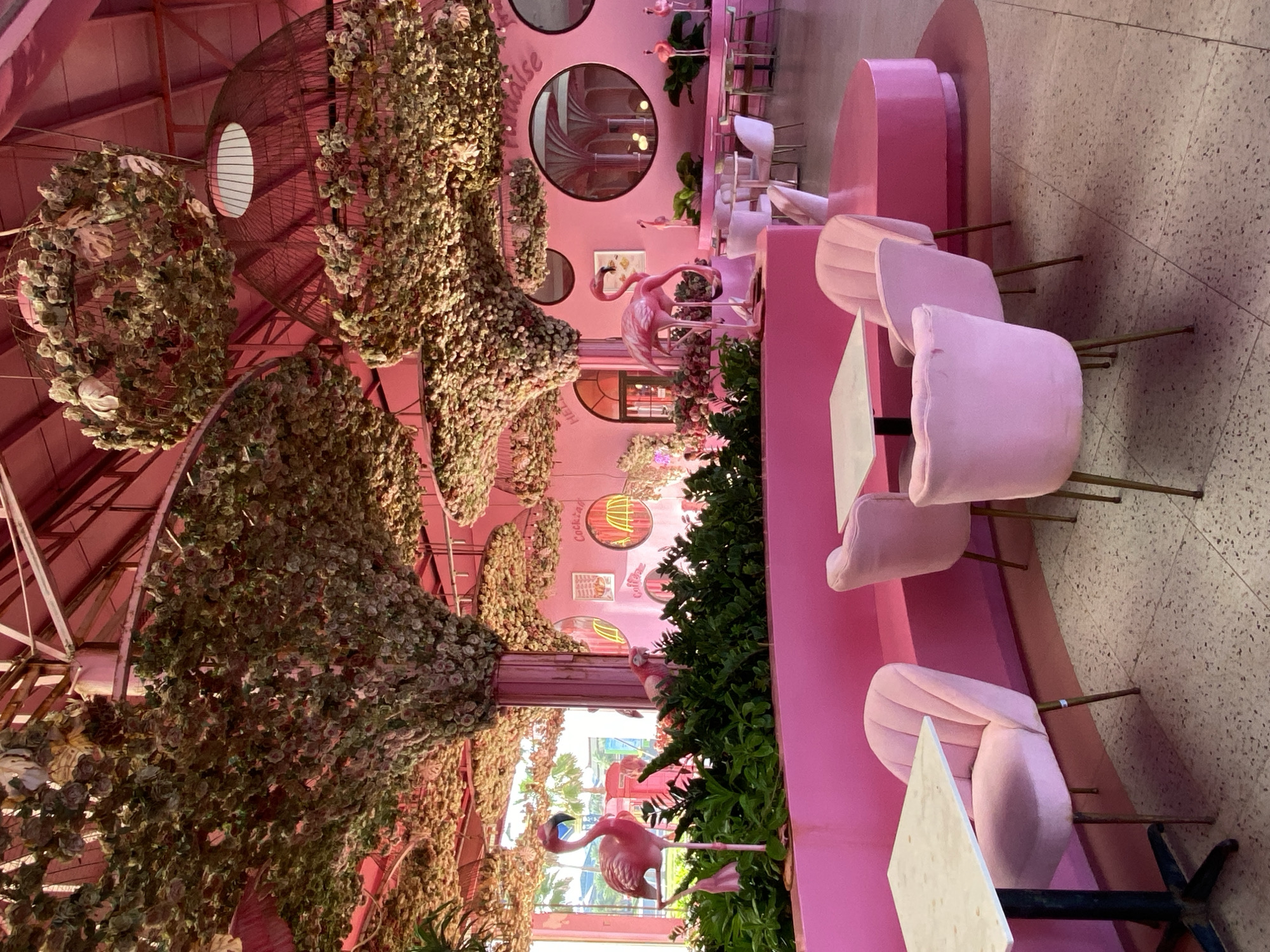The image depicts the interior of a brightly colored pink restaurant with a whimsical theme, possibly located in Tien Thanh Ward, Vietnam. The space is adorned with numerous floral decorations, with branches of artificial flowers cascading from the ceiling and framing the dining area. Several large, pink flamingo statues are strategically placed throughout the restaurant, adding to the playful ambiance.
The furniture consists of plush pink velvet chairs with gold-colored legs, surrounding white-topped tables with black bases. The seating arrangements suggest a dining or cafe setting. The walls are painted a vibrant pink and feature circular mirrored accents and framed artwork or signage, including a sign that reads "Cocktail." The floor appears to be a patterned terrazzo.
The overall impression is one of a highly stylized and themed establishment, designed to be visually appealing and perhaps aimed at a younger demographic or those seeking a unique dining experience. The lighting appears to be natural, coming from windows that are visible in the background, and augmented by artificial lights, some of which are integrated into the floral hanging decorations. No people are visible in the image, making it a still life of the restaurant's interior.