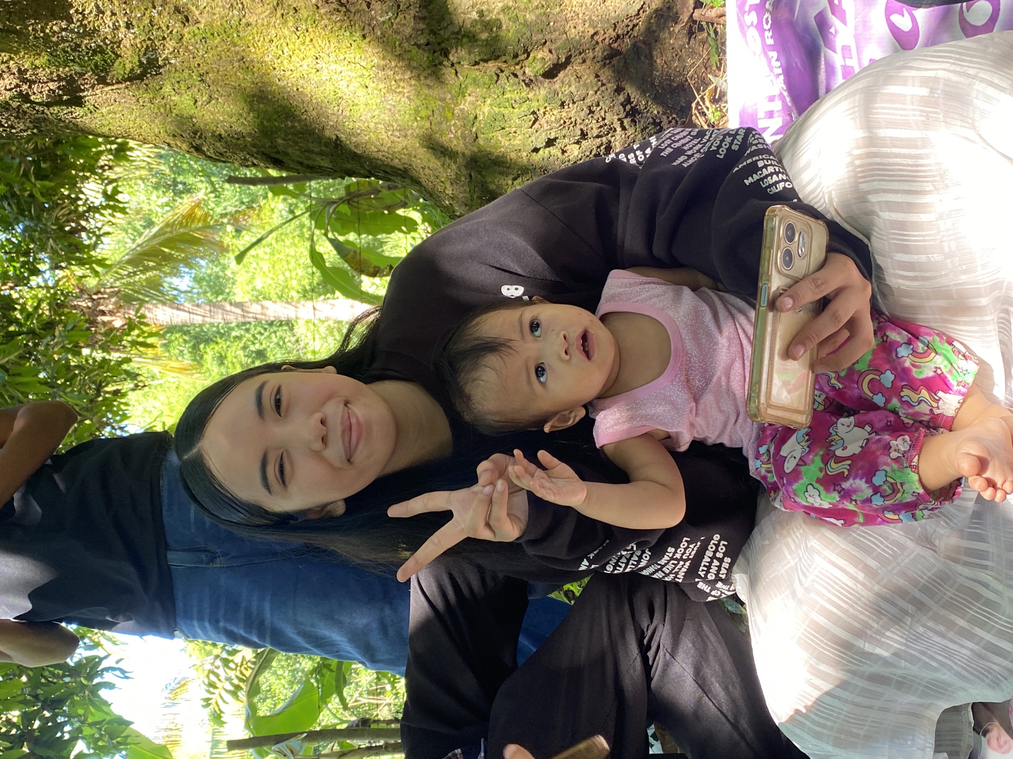The image captures a candid moment with two individuals and a young child outdoors, likely in a park or a rural area with lush vegetation.
In the foreground, a young woman is seated, holding a baby on her lap. The woman is wearing a black sweatshirt and a white, patterned skirt. She is looking towards the camera with a slight smile and her left hand is raised, forming a peace sign. The baby, dressed in a pink top and colorful pants, is looking up and to the side with an inquisitive expression. The woman is holding a gold-colored smartphone in her right hand.
Behind the woman, a person is standing, mostly obscured by the woman's head and shoulders. This person is wearing a black t-shirt and blue jeans. The background is filled with dense greenery, suggesting a natural, outdoor setting. A large tree with a textured, moss-covered trunk is visible on the right side of the frame, casting dappled sunlight on the scene. The overall impression is one of a relaxed, familial gathering in a natural environment.
While the image itself does not contain direct visual cues to definitively place it in Davao City, Philippines, the presence of tropical foliage and the general appearance of the people are consistent with a location in the Philippines. For example, the specific types of trees and the style of clothing worn by the individuals are common in tropical climates.