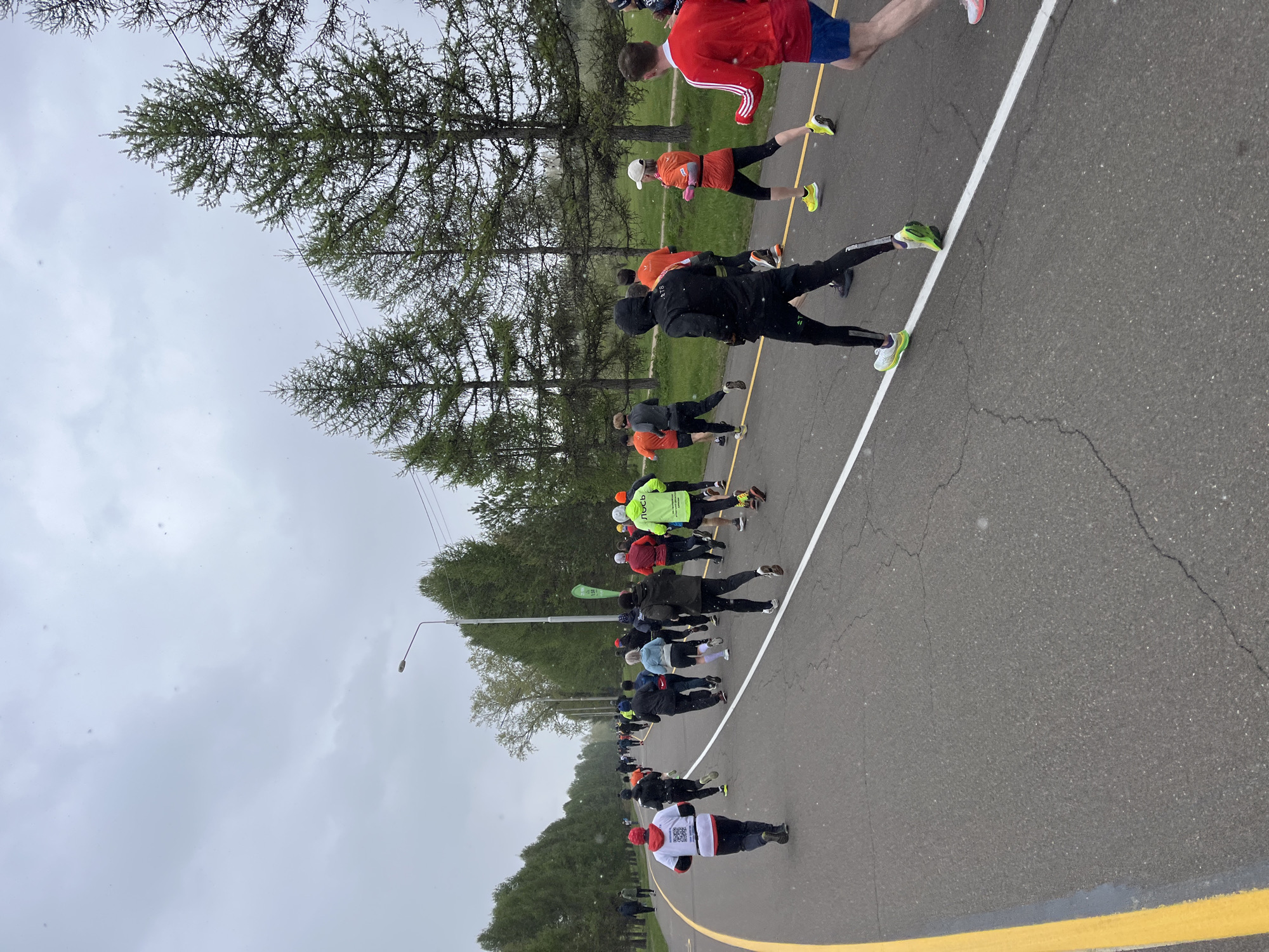 The image shows a road in Krasnoyarsk, Russia, with a group of people participating in a running event. The runners are of varying ages and are wearing different athletic attire. The road is paved with asphalt and marked with white and yellow lines. There are trees on either side of the road, and a cloudy sky is visible above.