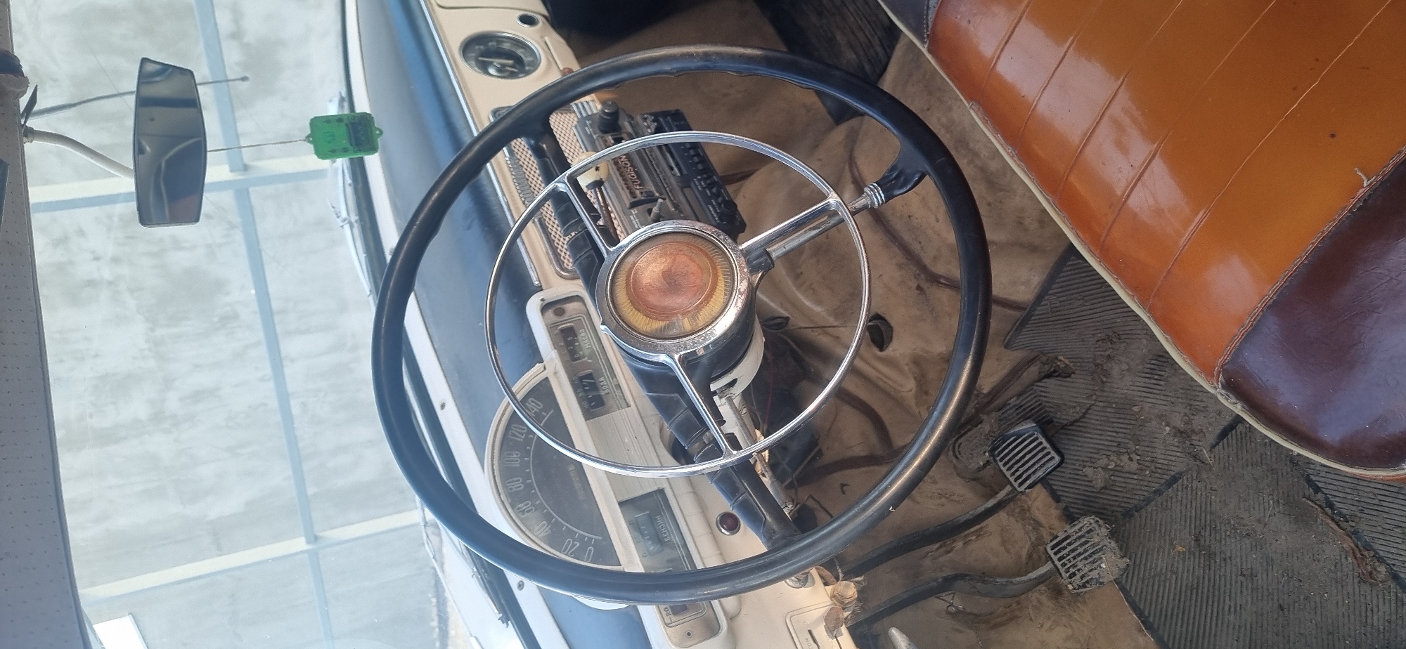 An interior view of an older car, with a black steering wheel featuring chrome spokes. The dashboard is white with a speedometer and other gauges. A radio is visible above the gauges. A rearview mirror is attached to the windshield, along with a green electrical component. The seat is a two-toned brown leather. The floor is dirty. The location is Ленинское сельское поселение, Ukraine.