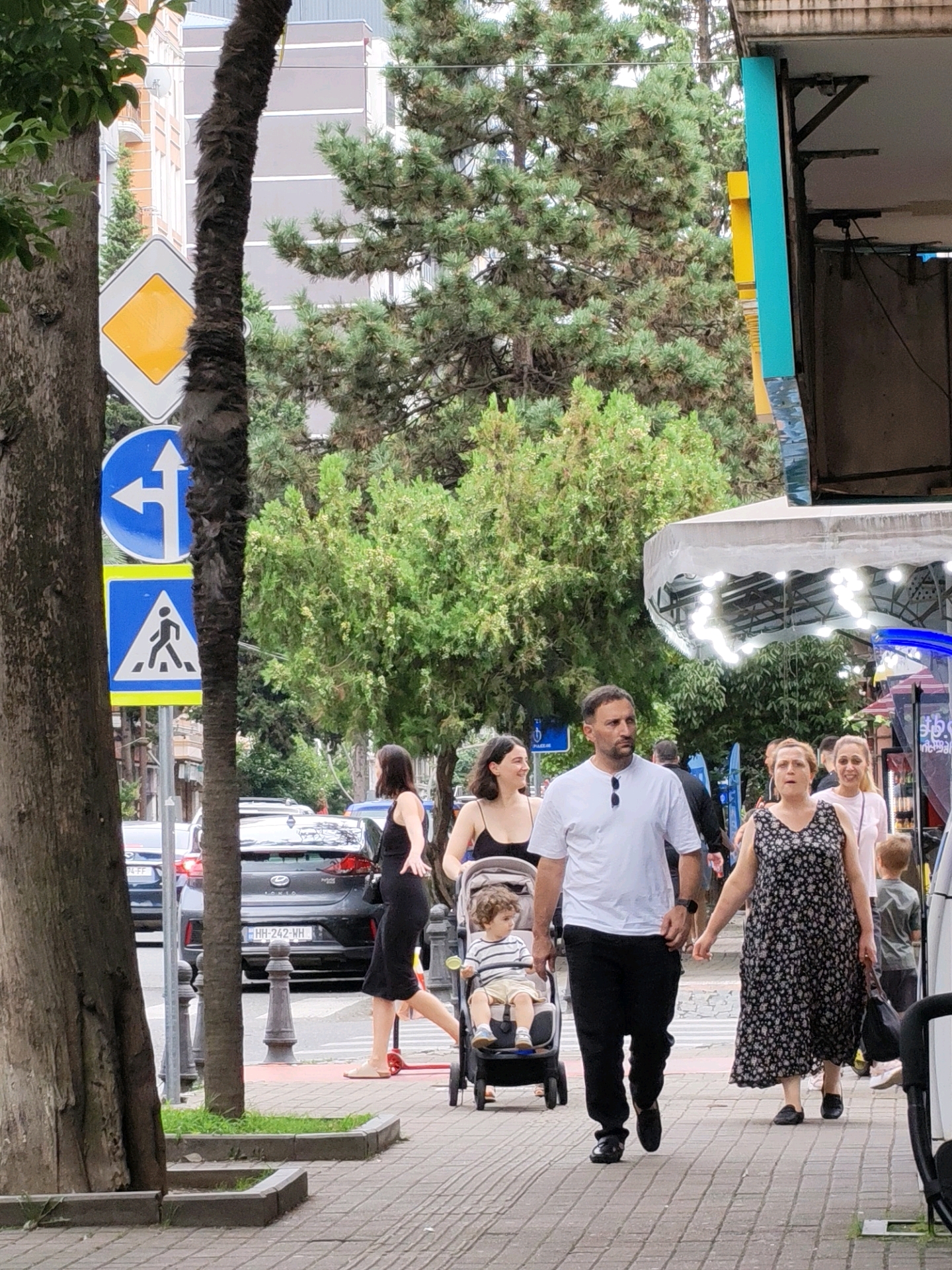 The image depicts a street scene in Batumi, Georgia, near the intersection of Rustaveli Avenue and Gorgasali Street, as indicated by the architecture and street signs. On the right, a pedestrian walkway is visible, where several people are walking. A family is present, with a man in a white t-shirt and black pants walking next to a woman in a black and white floral dress. They are holding hands. A child, around 3-4 years old, is in a stroller being pushed by someone behind them. To the left of the walkway, a busy street with cars is visible. Traffic signs, including a yield sign and a pedestrian crossing sign, are present on the side of the road. The background features buildings and lush greenery, characteristic of Batumi's urban landscape. The overall atmosphere appears to be a normal day in a bustling city, with people going about their daily activities.