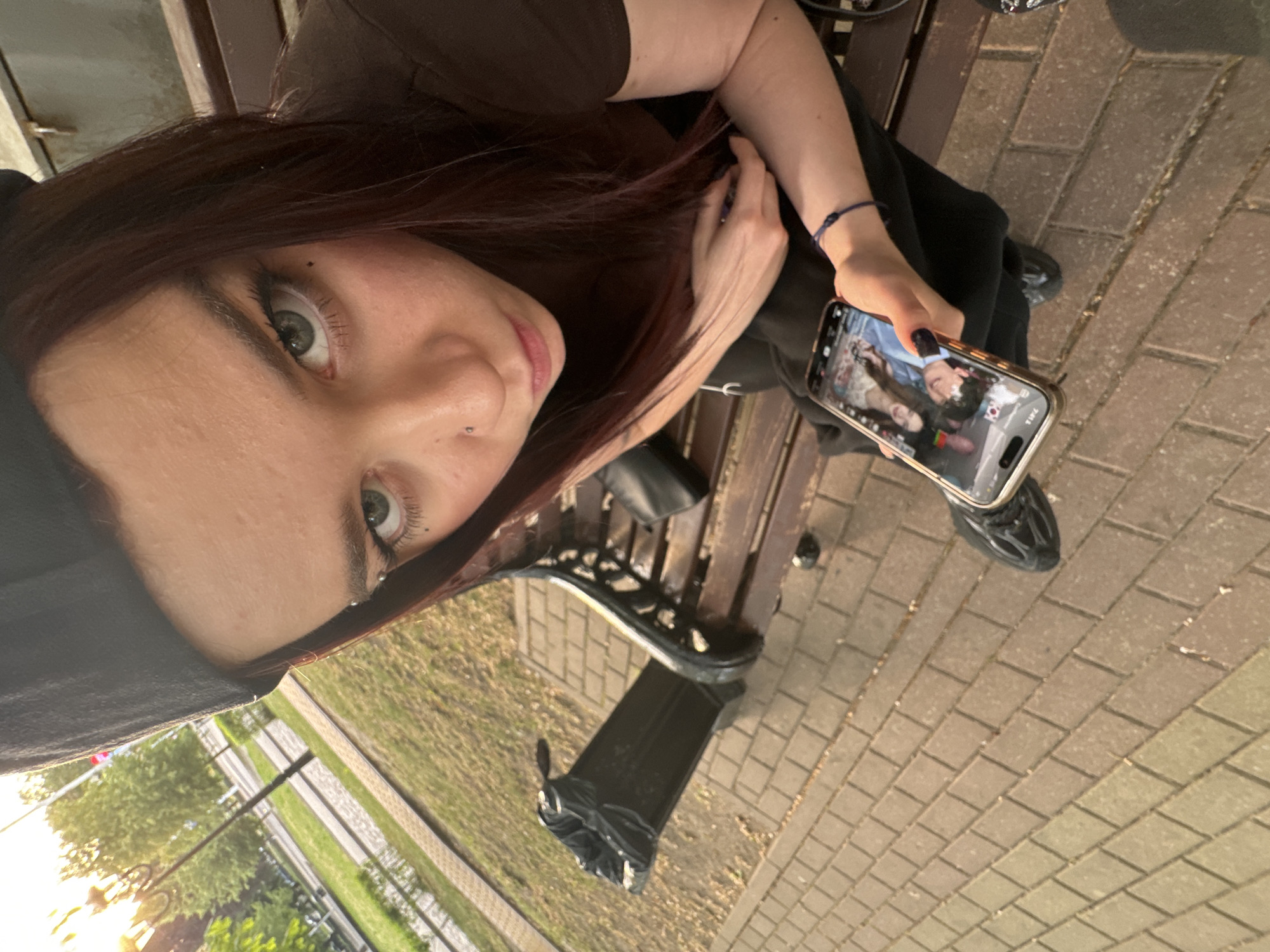 A selfie taken by a person with auburn hair, light skin, and a nose piercing. They are wearing a black hat and a brown shirt. The person is sitting on a wooden bench in a park with a paved walkway. They are holding a phone displaying an image of a couple. A trash can is visible next to the bench. The setting appears to be a city park, with trees and a green lawn in the background.