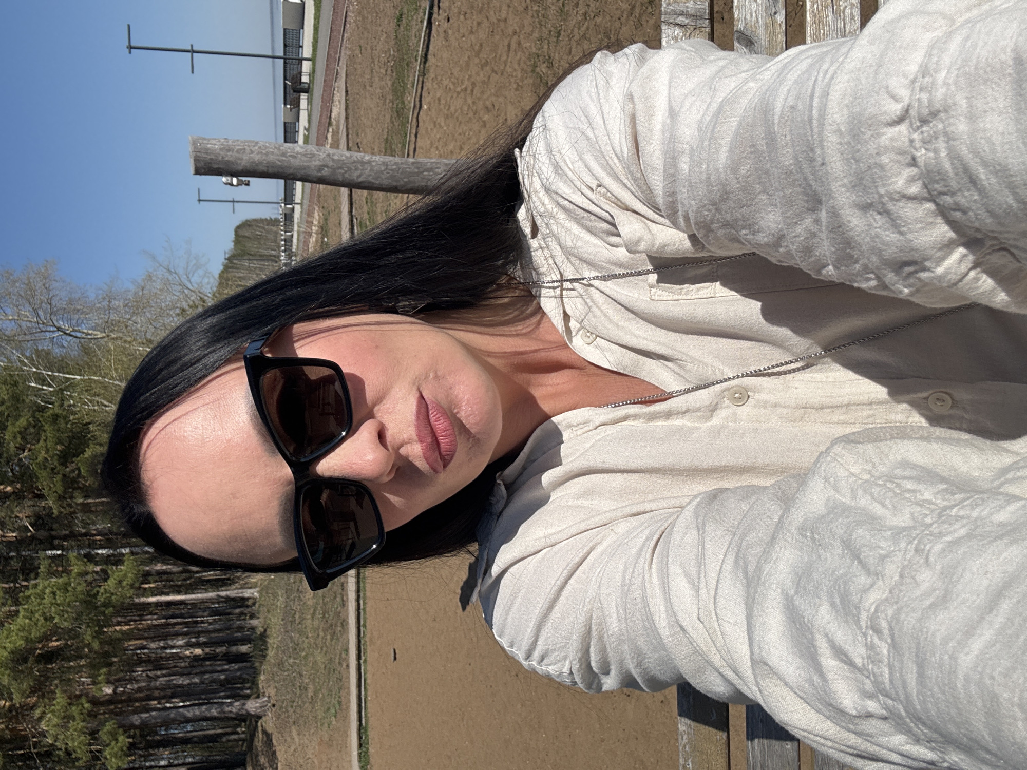A woman sits on a wooden bench in Chaikovsky, Russia. She wears dark sunglasses and a light-colored button-down shirt. Her long, black hair contrasts with the shirt. In the background, trees and a body of water are visible under a clear blue sky. A wooden post is located near her. The ground is mostly covered in dirt.