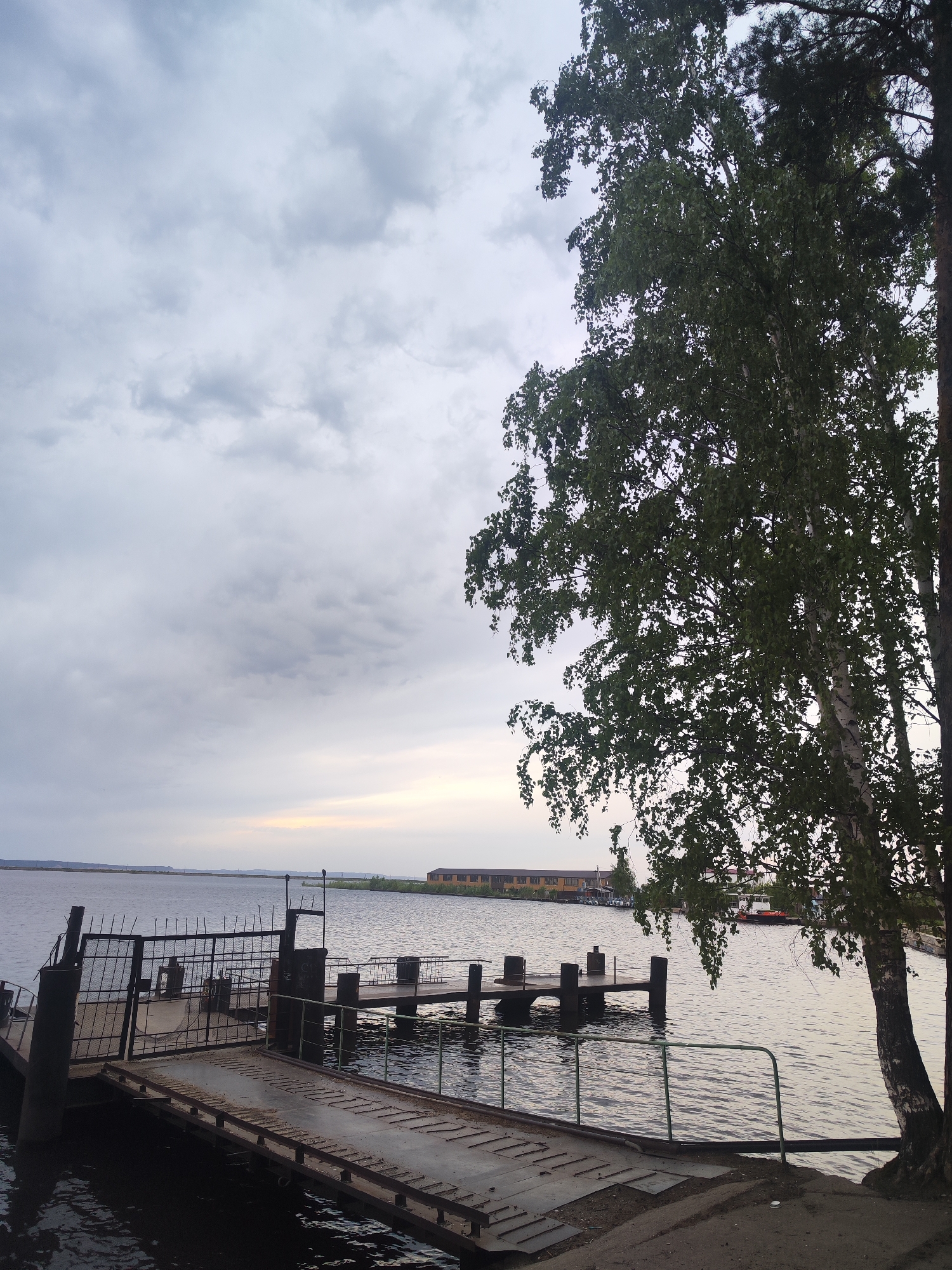 The image captures a waterfront scene in Unknown, Unknown, featuring a pier extending into a body of water. The pier is constructed of metal and wood, showing signs of wear and age. A ramp connects the pier to the land. To the right, a tall tree with dense foliage partially obscures the view. In the background, across the water, are long, low buildings. The sky is overcast with a mix of gray and white clouds. The overall lighting is dim.