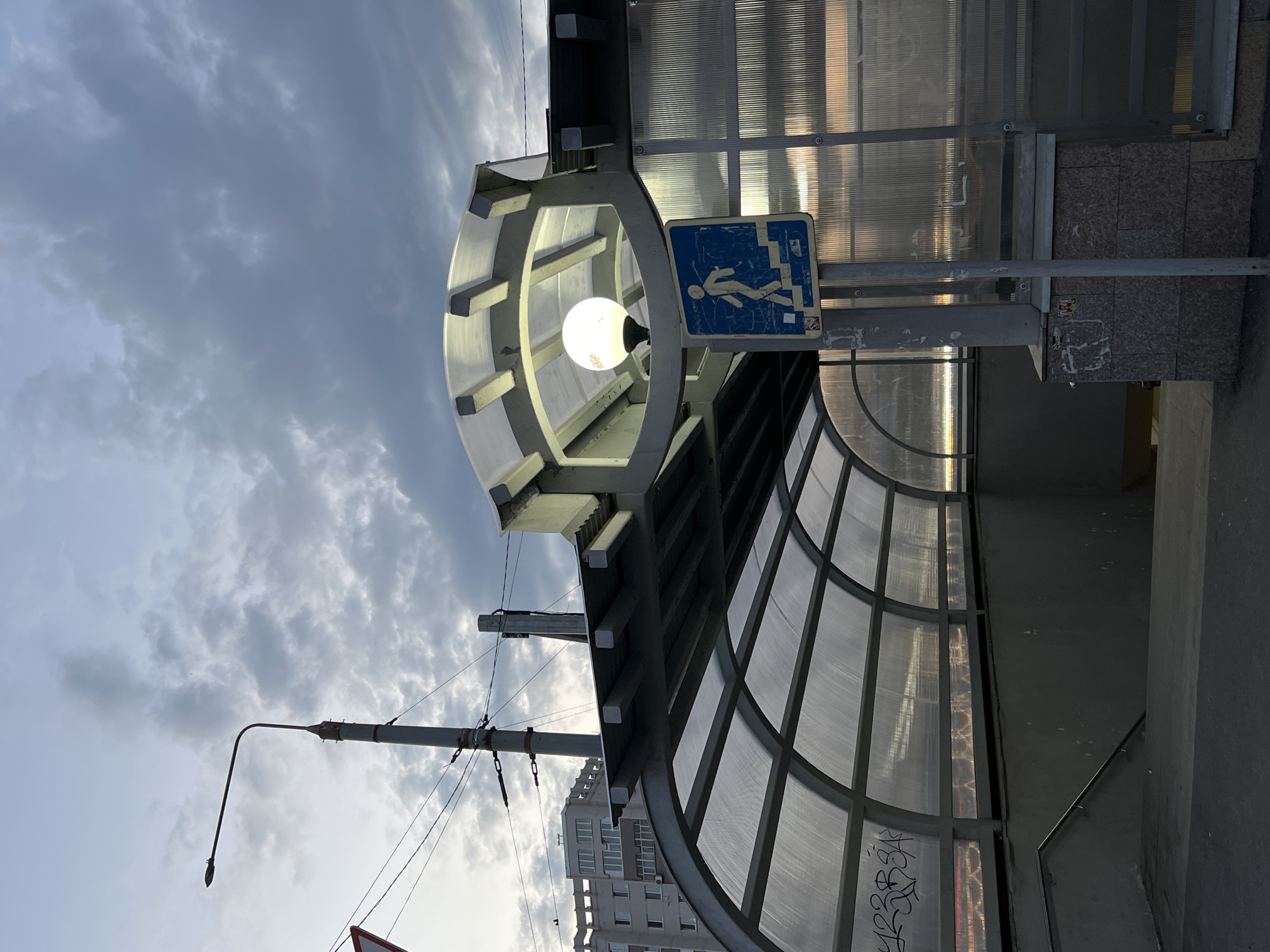 The image shows an overhead view of a pedestrian underpass in Kazan, Russia, during twilight. The underpass is covered by a translucent, arched roof made of polycarbonate panels supported by a metal frame. The interior of the underpass appears to be descending, with concrete stairs and a metal railing visible. On the right side of the entrance, a blue pedestrian crossing sign with a white silhouette of a person walking down stairs is mounted on a pole. Above the entrance, a street lamp with a spherical bulb is illuminated, casting a warm light. The sky is cloudy and overcast, with a hint of sunset colors reflecting on the translucent roof. In the background, a multi-story residential building is visible, along with a street lamp and overhead wires. The overall impression is a utilitarian urban scene at dusk.