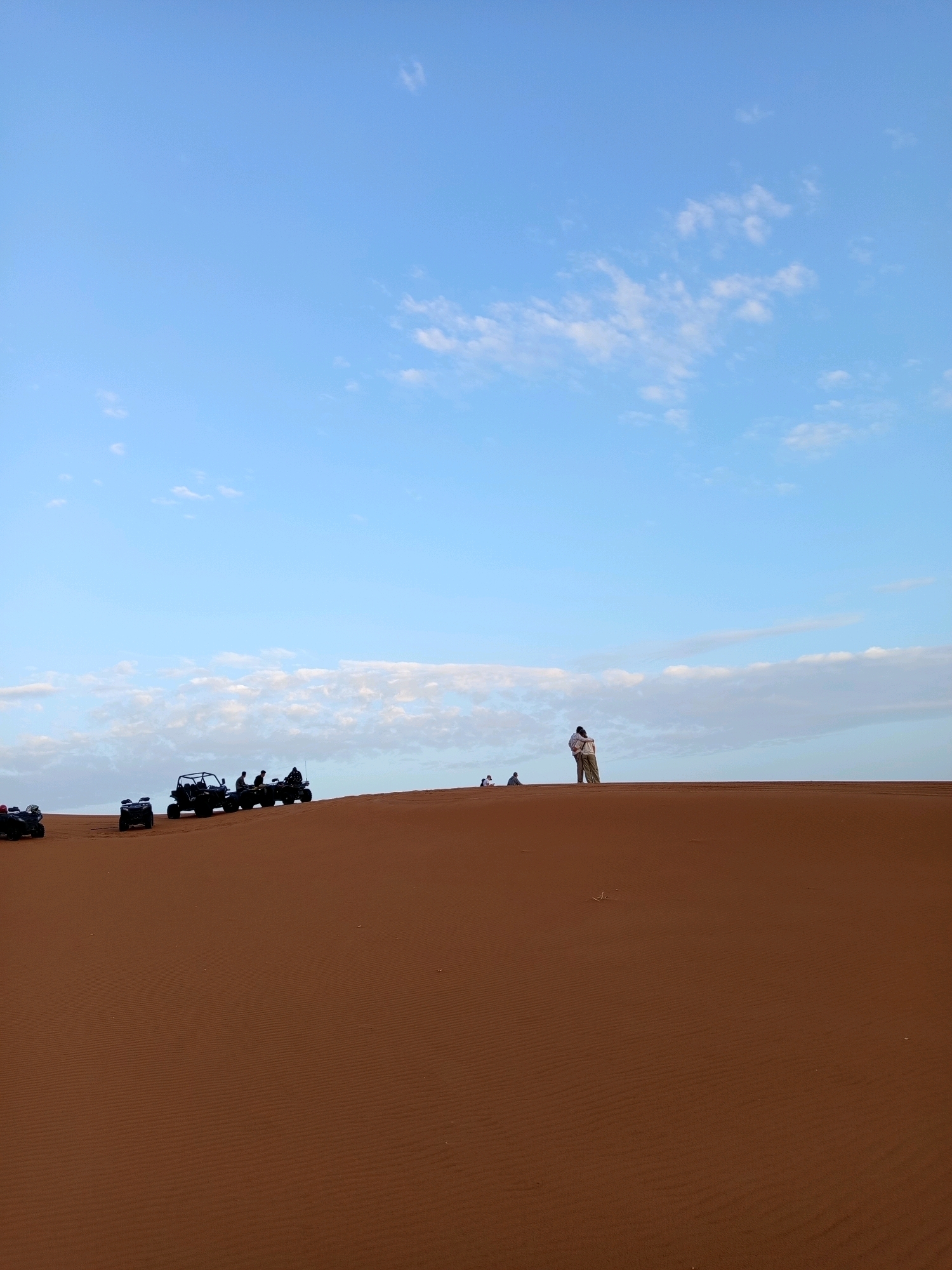 The image depicts a desert landscape in Rissani, Morocco, characterized by undulating sand dunes. The sky above is a clear blue with scattered white clouds. Several all-terrain vehicles (ATVs) are present on the crest of a dune. In the foreground, one ATV is partially visible on the left. Further up the dune, a line of ATVs are parked, with silhouettes of people visible on or near them. On the right side of the dune, two individuals are standing together, one embracing the other. Two smaller figures are seated in the distance on the left. The sand exhibits the characteristic rippled texture of a desert environment.
