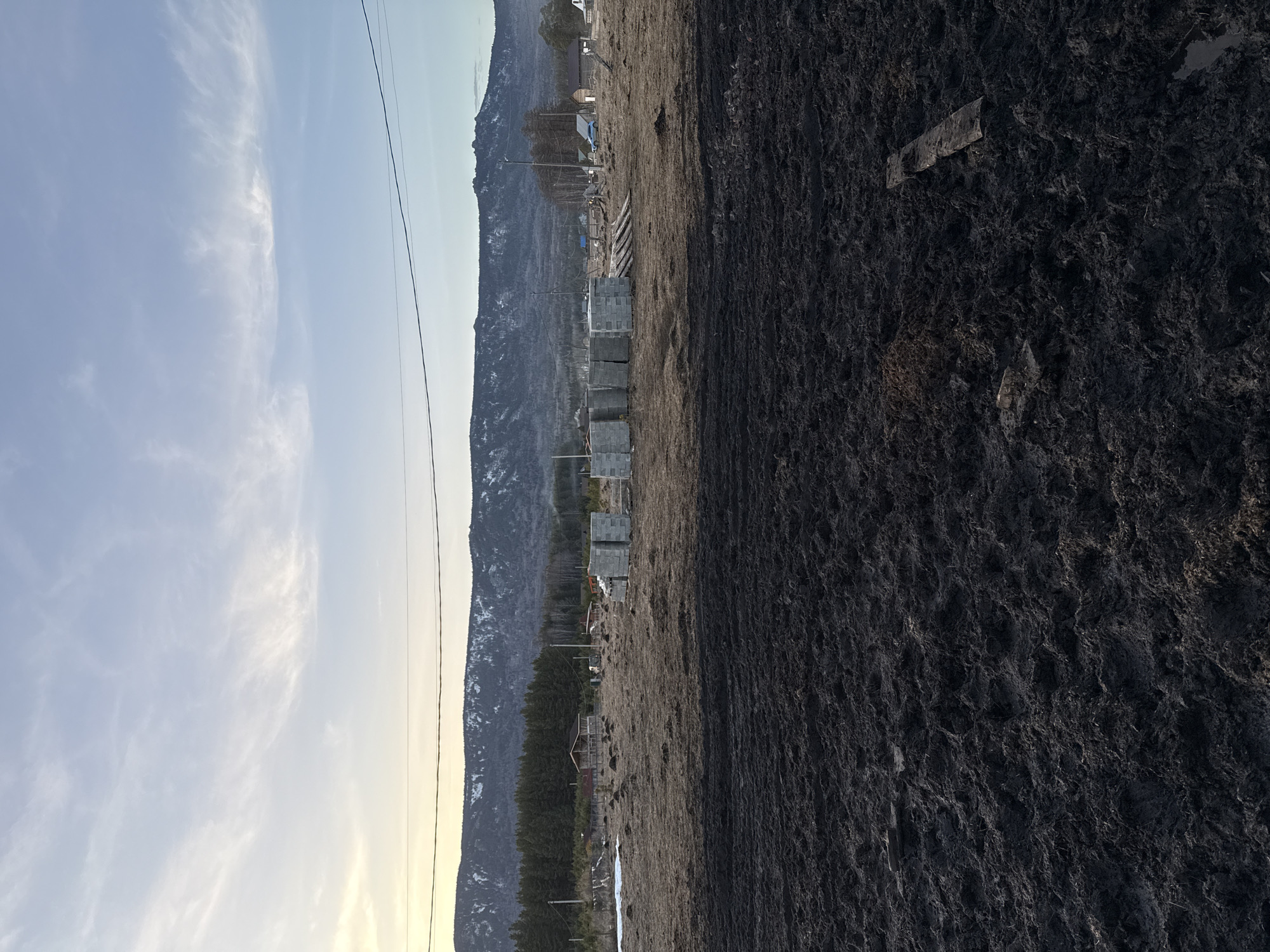 The image depicts an open area in Tyulyuk, Russia, under what appears to be either early morning or late afternoon light. The foreground consists of dark, churned earth heavily saturated with mud, exhibiting a rough texture with visible indentations and a small puddle on the far right. Two weathered wooden planks are partially submerged in the mud on the right side of the frame.
Moving towards the midground, the terrain transitions to lighter brown, dry, sparse grass with scattered patches of darker soil and low mounds. In this area, multiple organized stacks of light gray concrete blocks are present, positioned in several rows. Near these blocks, a loose pile of wooden planks or lumber is visible. Further back, rudimentary wooden buildings or structures are dispersed across the landscape, including one with a darker roof on the left and a more developed structure on the right. A light blue sedan and a white pickup truck are discernible parked near the structures on the right. Several utility poles with overhead power lines extend horizontally across the scene. A dense line of dark green coniferous trees forms a prominent backdrop for the immediate settlement area.
The background is dominated by a range of snow-capped mountains under a pale blue sky. Wispy white clouds are scattered across the sky, with a yellowish hue near the horizon on the left side, indicative of low sun angle. No individuals are visible within the scene.