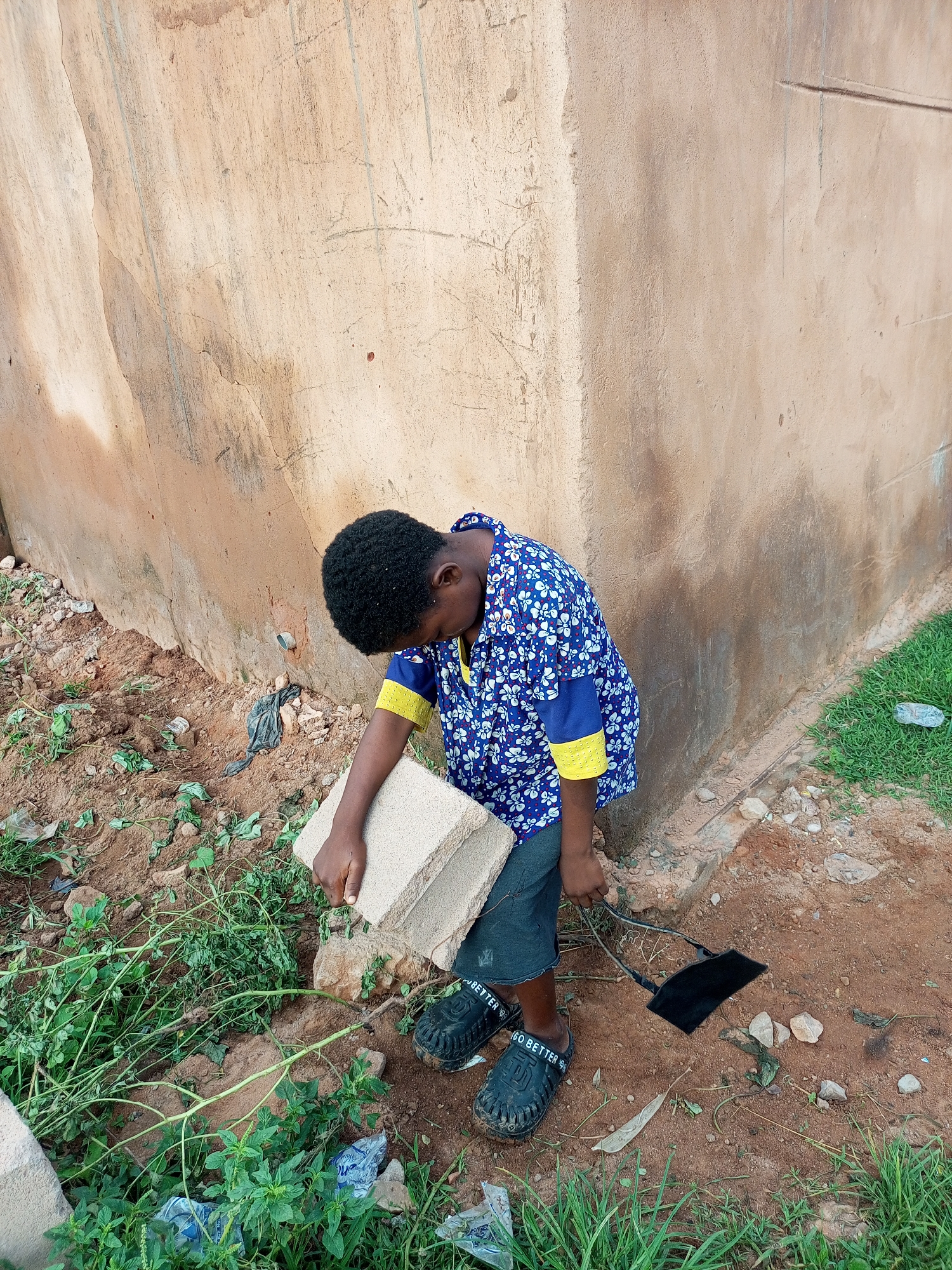A young African child, dressed in a blue and white floral patterned shirt with yellow accents and dark shorts, stands outside next to a rough, textured, beige-colored wall. The child is looking down, holding a light-colored rectangular object in their right hand. A black cord with a small black object attached is visible on the ground near the child's feet. The ground is covered with reddish-brown soil, scattered rocks, and patches of green grass and vegetation. The overall setting appears to be an outdoor area, possibly in a less developed part of Tempure, Nigeria, with a utilitarian or unfinished construction element suggested by the wall.