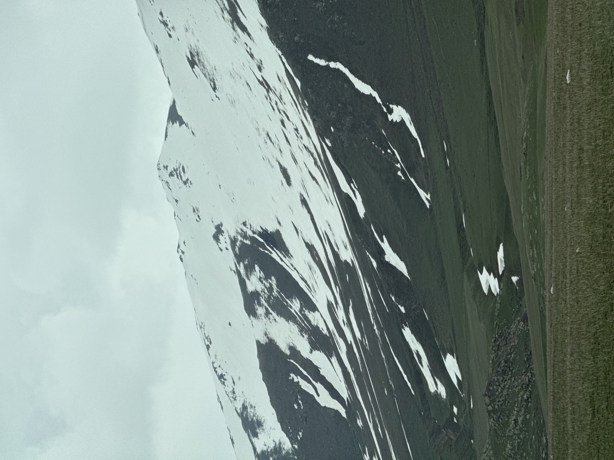 The image depicts a mountain range with snow-covered peaks, possibly located in the vicinity of Aparan, Armenia. The landscape is characterized by a mix of rocky terrain, snow patches, and grassy slopes. The sky is overcast, suggesting a cloudy or foggy weather condition. There are no discernible people, subjects, objects, or interactions apparent in the image other than the natural elements of the environment. The scene is dominated by the mountainous terrain and its various features.